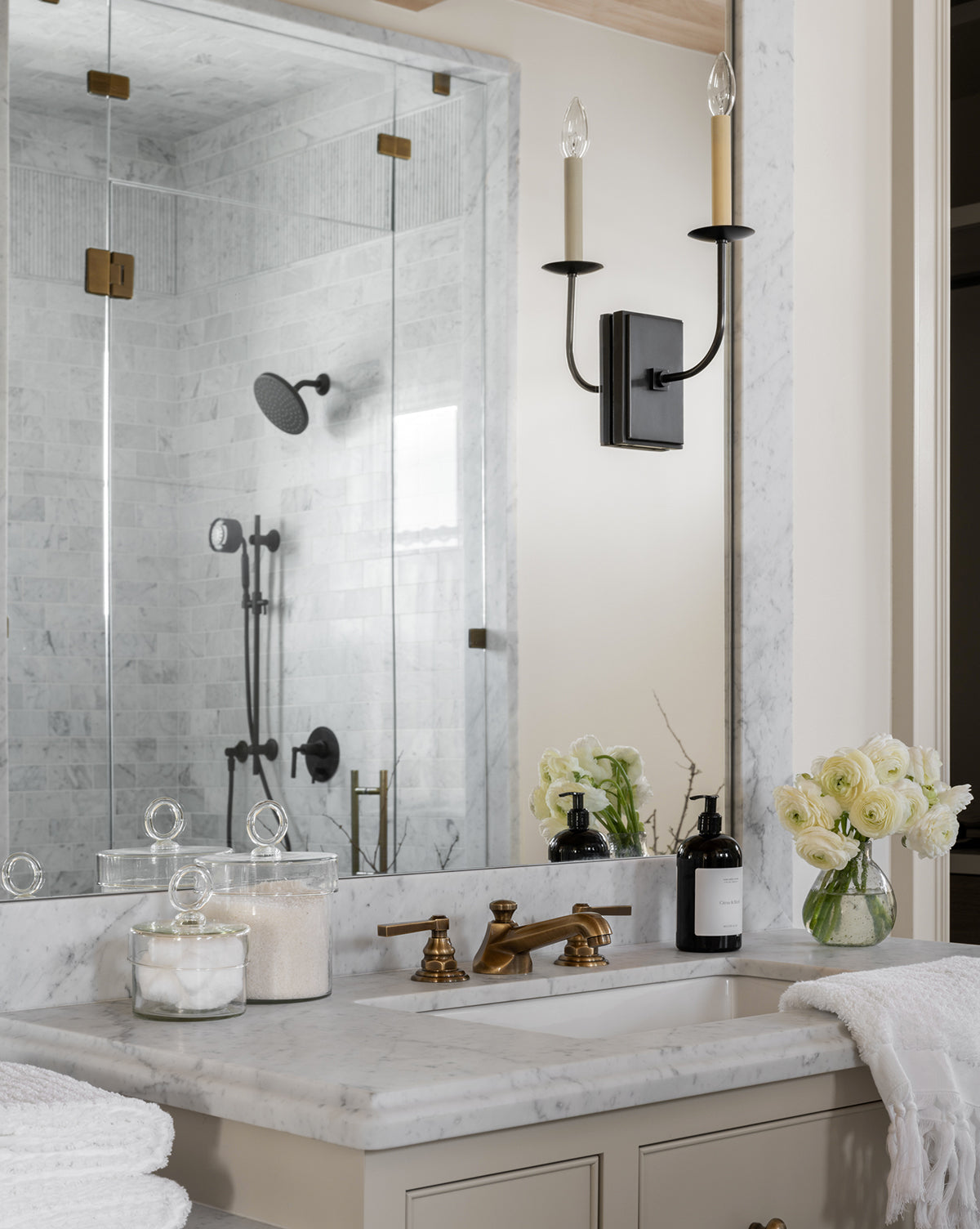 Elegant bathroom featuring a marble countertop, brass faucet, the Glass Lidded Canister with a round handle, soap dispenser, fresh white flowers in a vase, and a mirrored wall reflecting a glass-enclosed shower with black fixtures.