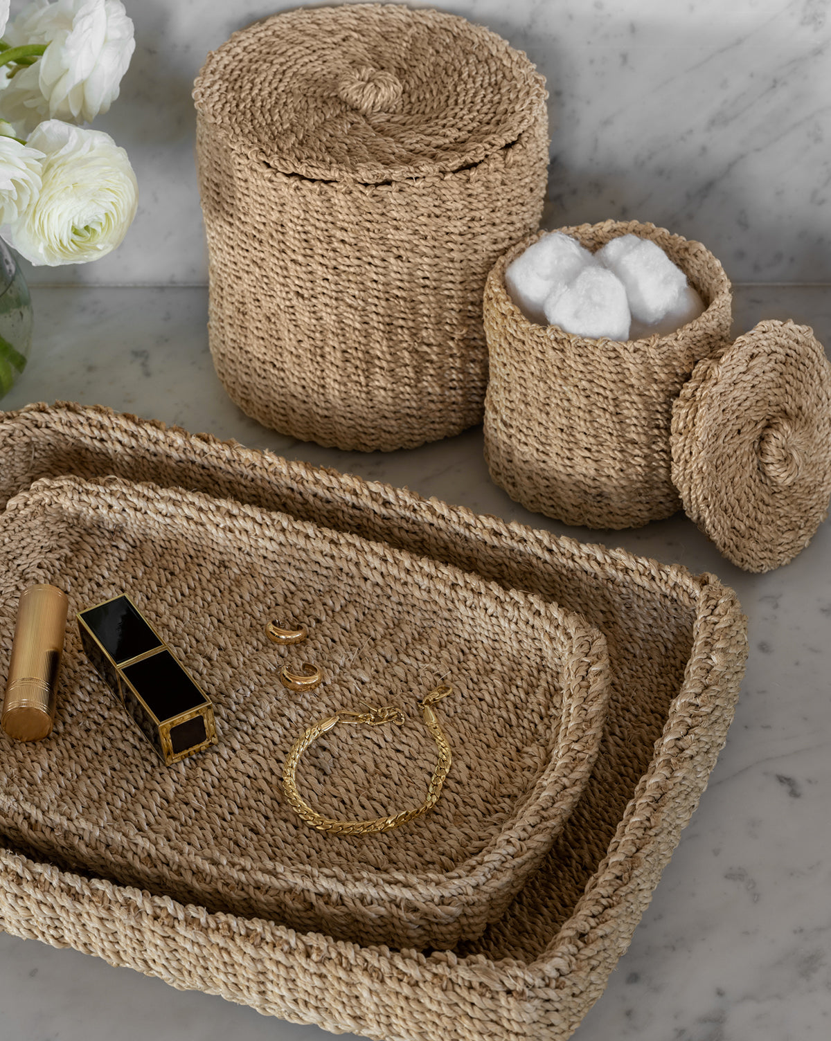 The Deryn Woven Tray holds gold jewelry and lipstick on a marble surface beside two woven baskets, one open and filled with cotton balls. White flowers appear in the corner.