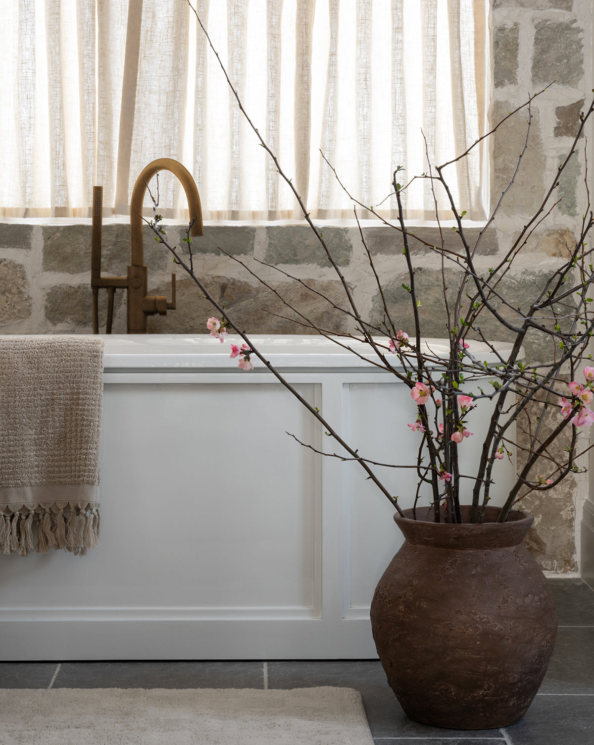 A rustic bathroom featuring a white bathtub, beige towel, bronze faucet, and the Demetria Terracotta Vase holding pink-blossomed branches; stone wall and sheer curtains serve as the backdrop.