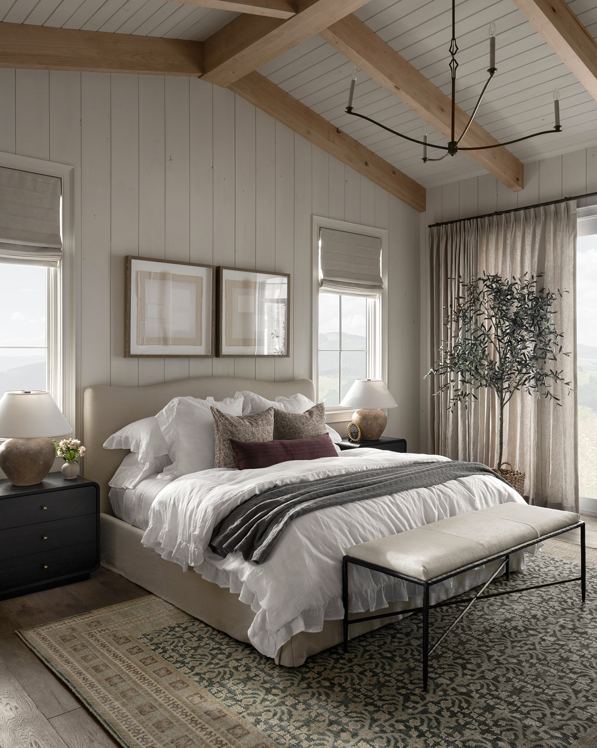 A cozy, modern bedroom with a large bed dressed in the Ruffle White Linen Duvet Cover, dark nightstands, table lamps, an upholstered bench, framed art above the bed, an area rug, a potted tree, and large windows with light curtains.