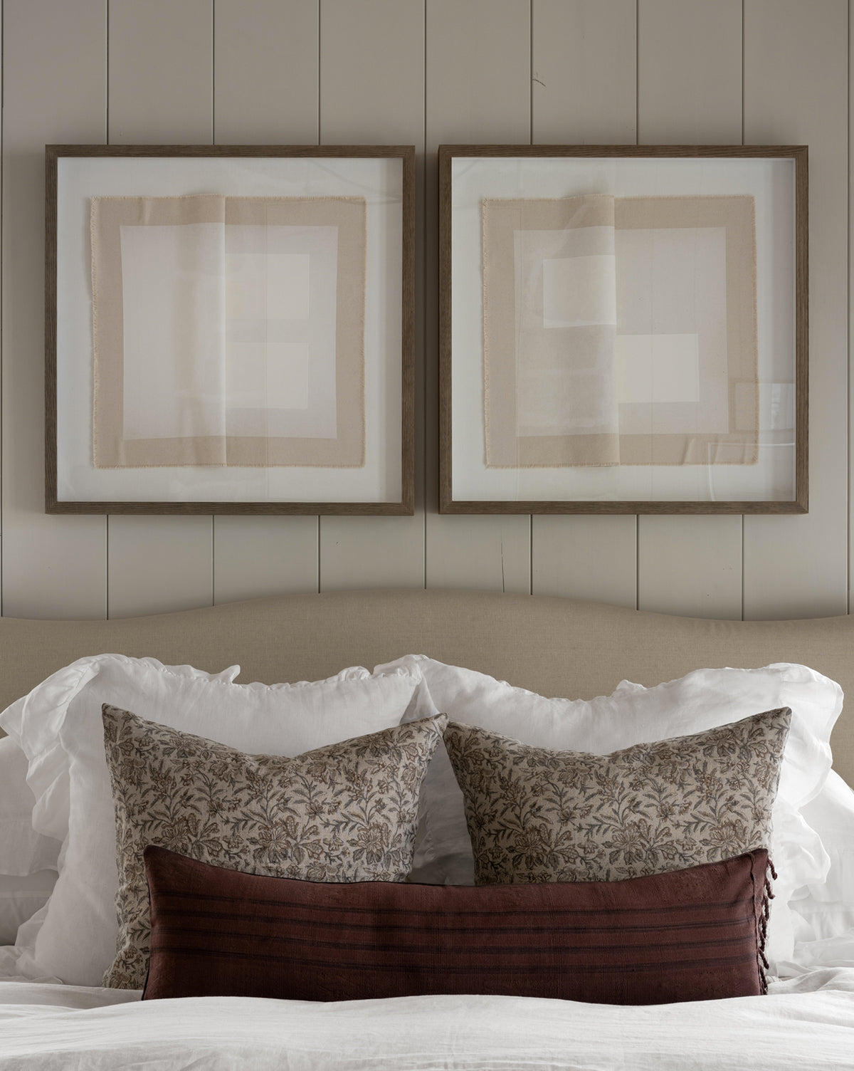 A neatly made bed with white bedding, two patterned pillows, a dark brown lumbar pillow, Ruffle White Linen Shams (Set of 2), and two abstract framed artworks on a light-colored shiplap wall above the headboard.