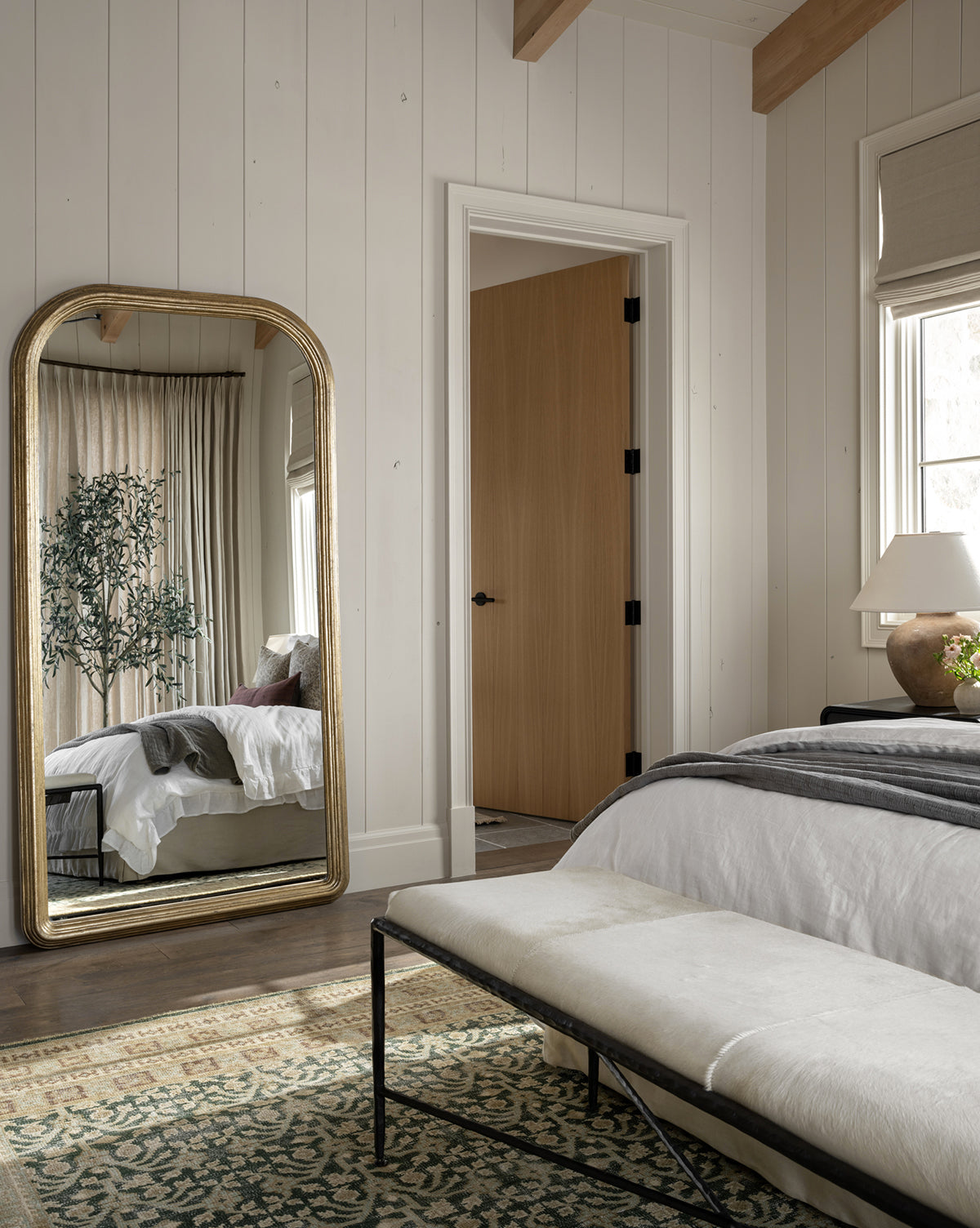 A cozy bedroom with light wood walls features the Giada Floor Mirror with an elegant frame, reflecting a bed with white and gray bedding, a potted plant, a bench at the foot of the bed, and natural light through beige curtains, by McGee & Co.