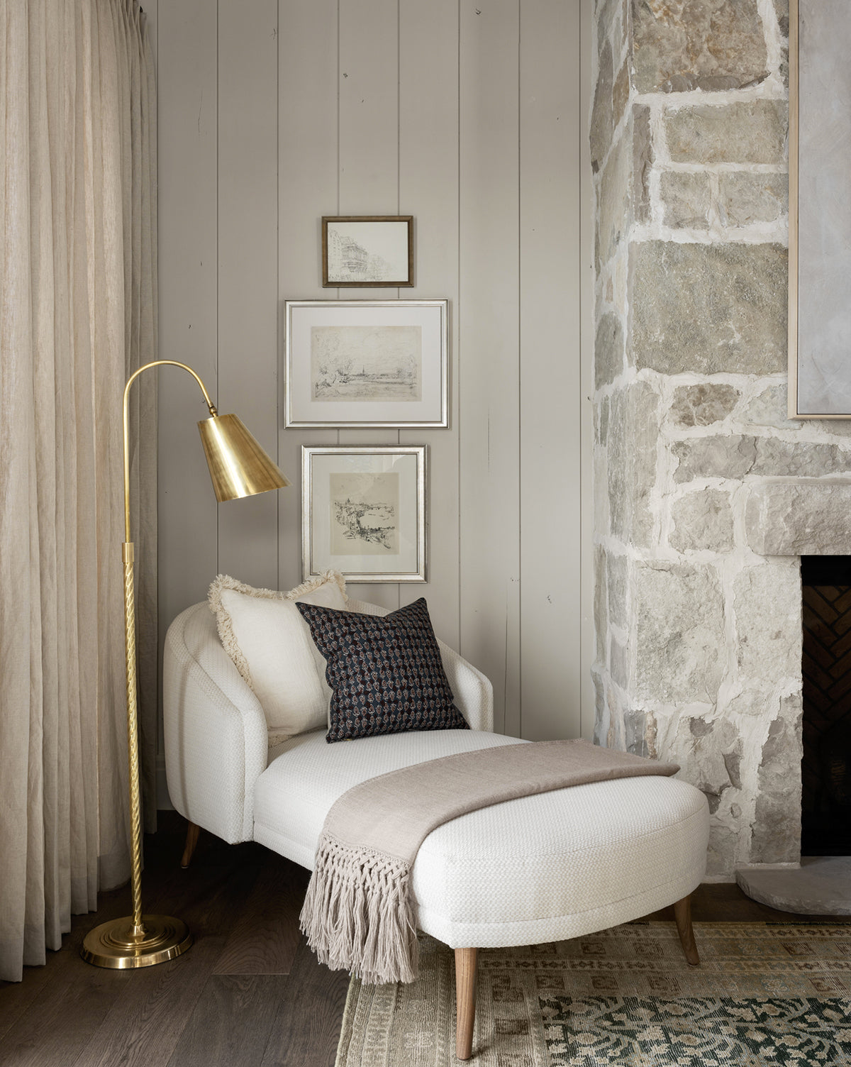 A cozy reading nook featuring a white chaise lounge, beige throw and cushion, neutral drapes, framed art on paneled walls, a stone fireplace, and elegant warmth from the Saylor Floor Lamp, by McGee & Co.