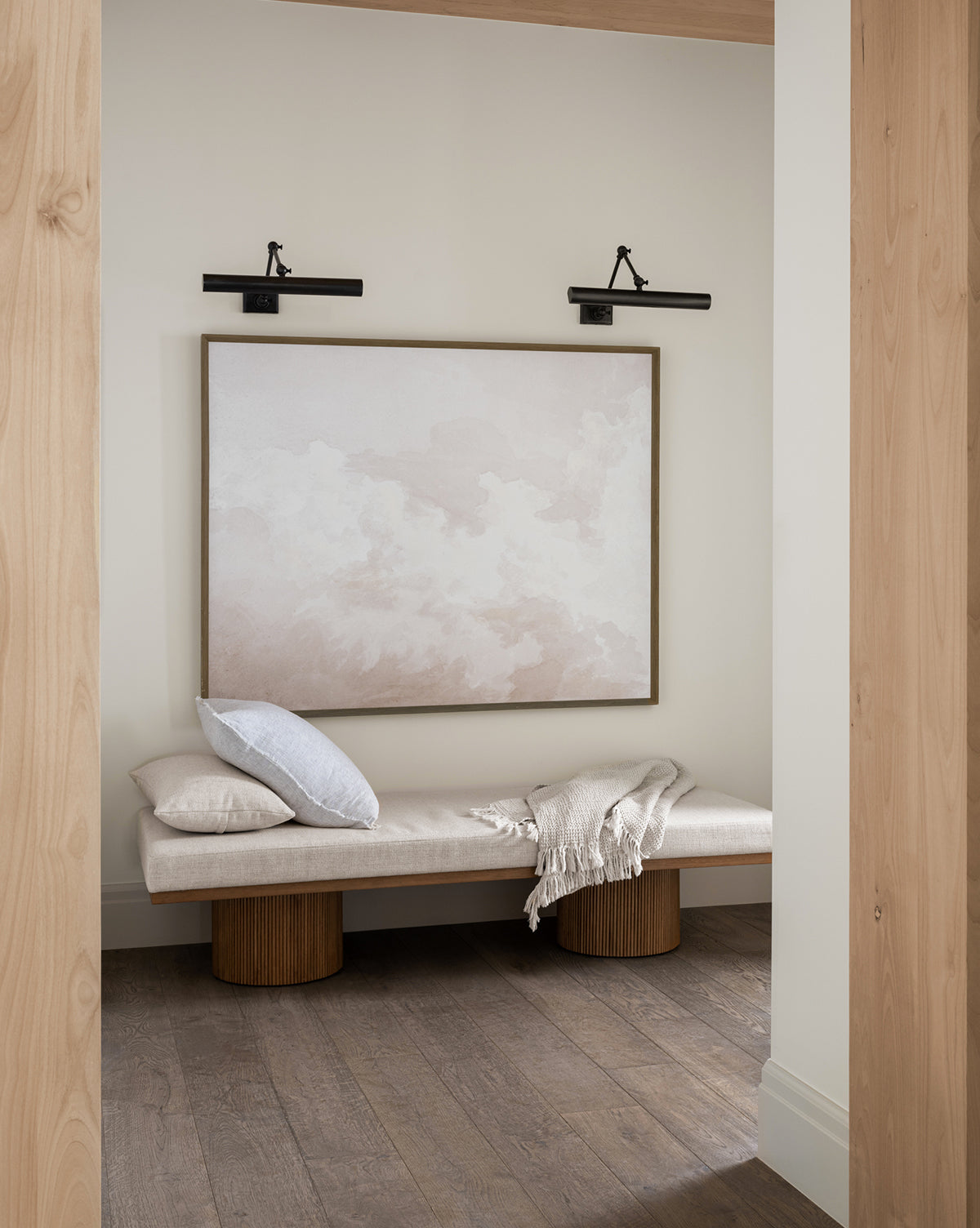 A minimalist entryway features a light wood bench with a pale cushion, white throw blanket, and the Meditation on Clouds artwork in a walnut frame above, illuminated by black wall lights. The space includes light walls and dark wooden flooring.