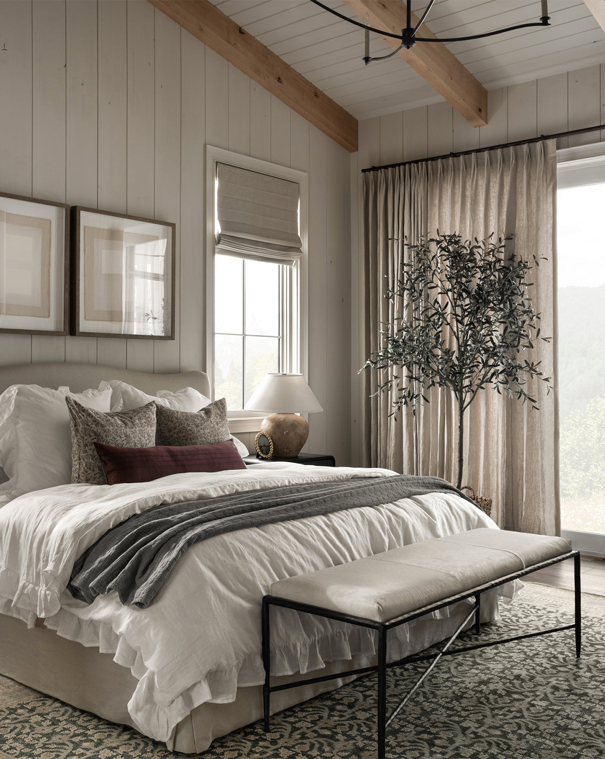 A cozy bedroom features a neatly made bed, neutral bedding, a decorative bench, large windows with light curtains, framed art, and the Faux 96 Olive Tree with Woven Pot adding natural charm to the wooden floor rug.