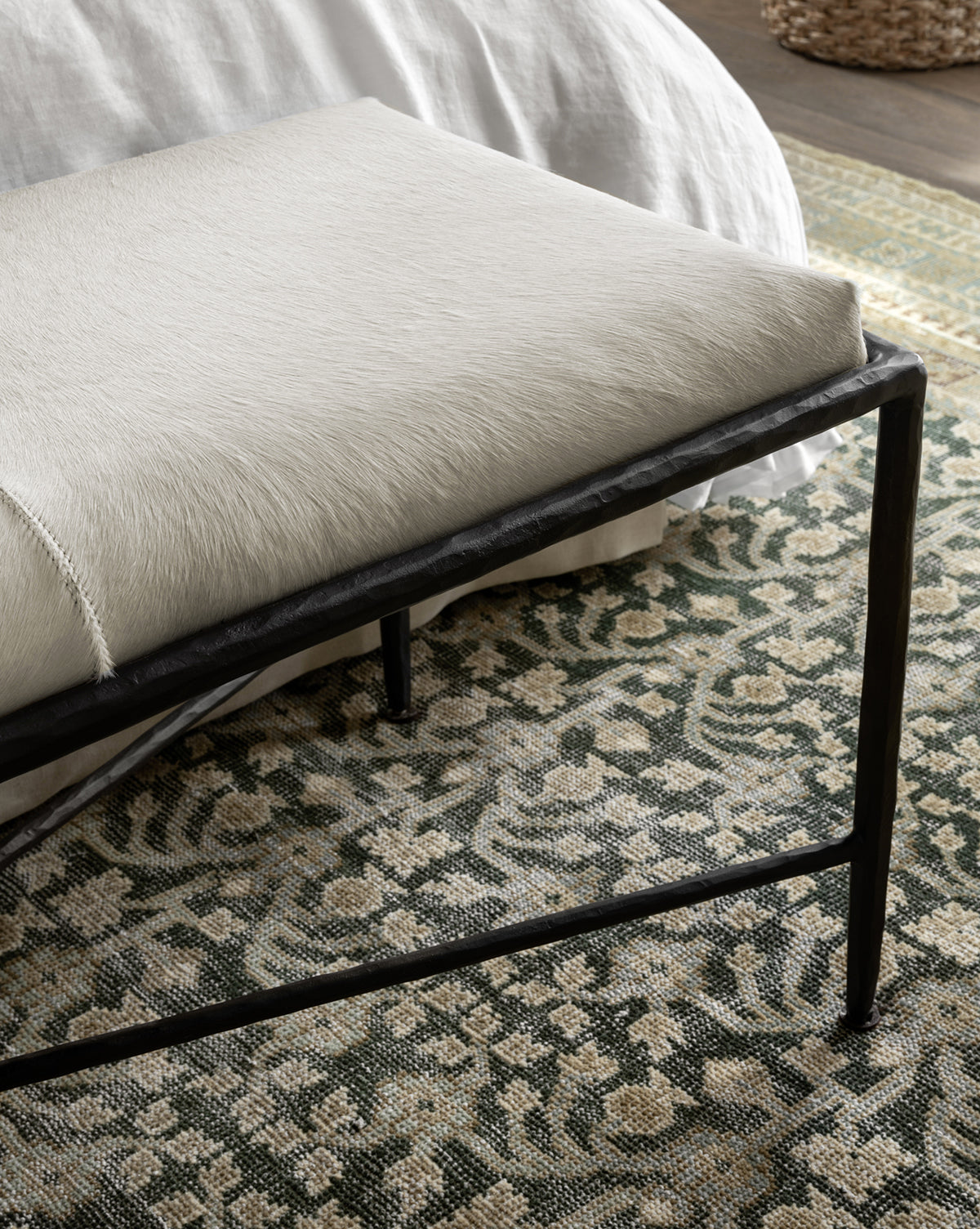 A close-up of a bench with a cream-colored upholstered seat and black metal legs, placed on the Evangeline Hand-Knotted Wool Rug with floral designs near a white bedspread, by McGee & Co.