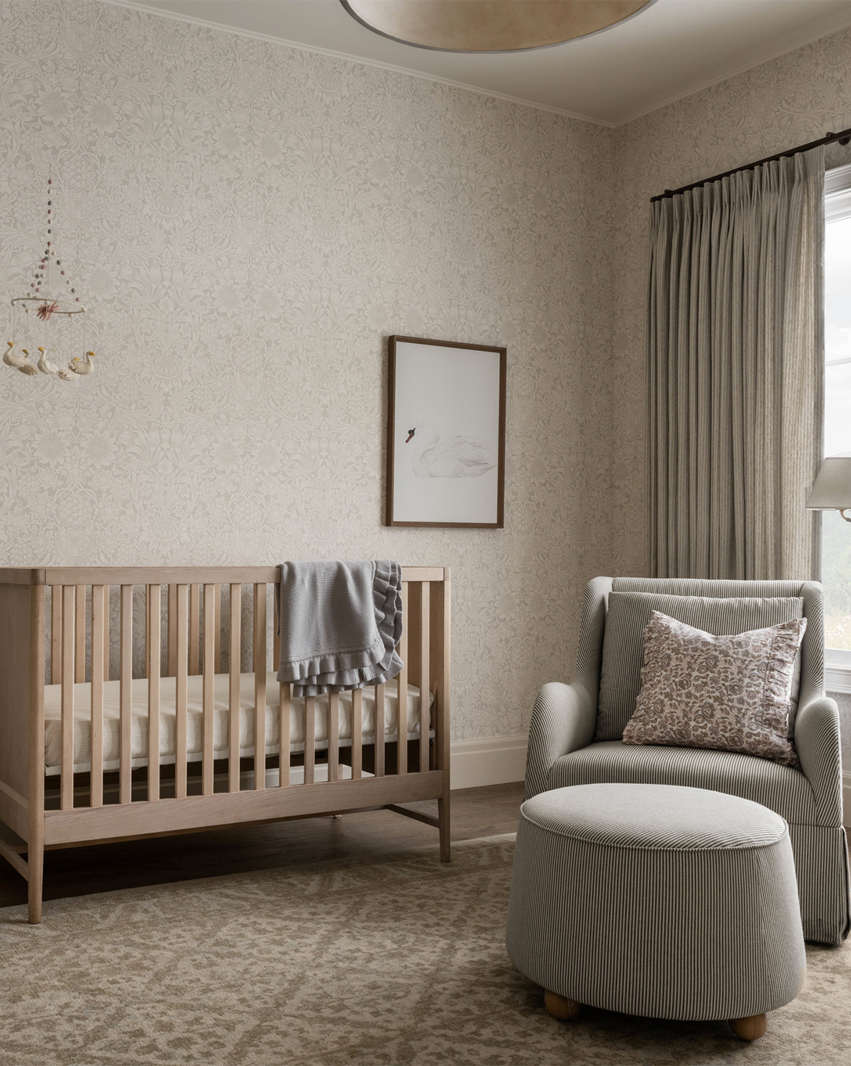 A cozy, neutral nursery features a wooden crib, gray blanket, armchair with patterned pillow, round ottoman, soft rug, long curtains, and the Neutral Swan framed wall art in walnut wood grain beside a swan mobile.