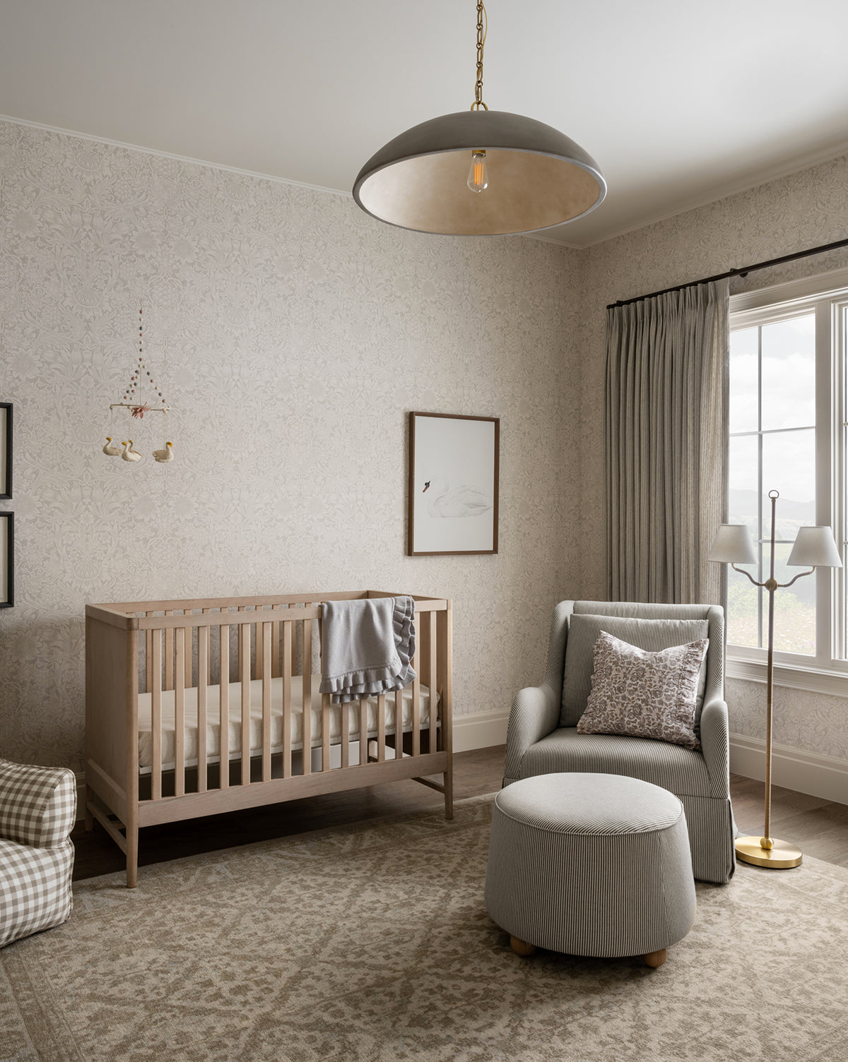 A cozy nursery featuring a light wood crib, gray armchair with ottoman, neutral patterned rug, soft curtains, swan wall art, animal mobile, jute rope accents, the Sirocco Floor Lamp, and plenty of natural light from a large window, by McGee & Co.