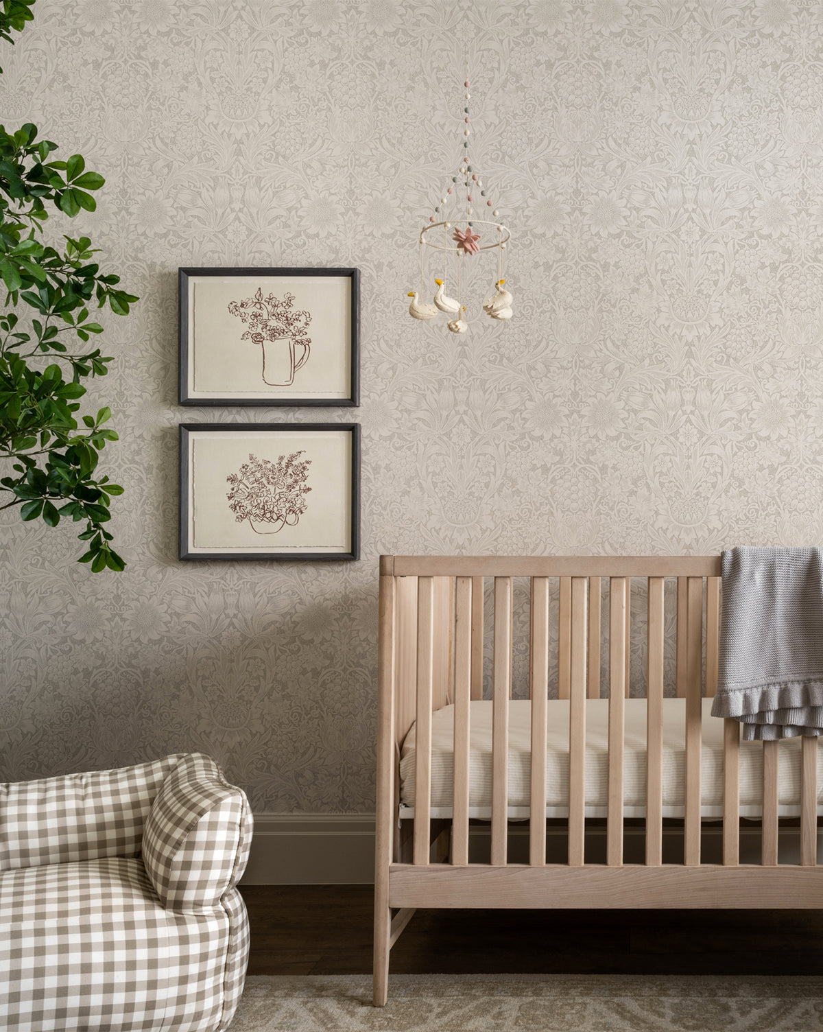 A cozy nursery features a light wood crib with the Easton Crib Sheet, a gray blanket draped over the rail, a plaid armchair, two framed botanical prints on the wall, a leafy green plant, and a sweet bird mobile hanging above.