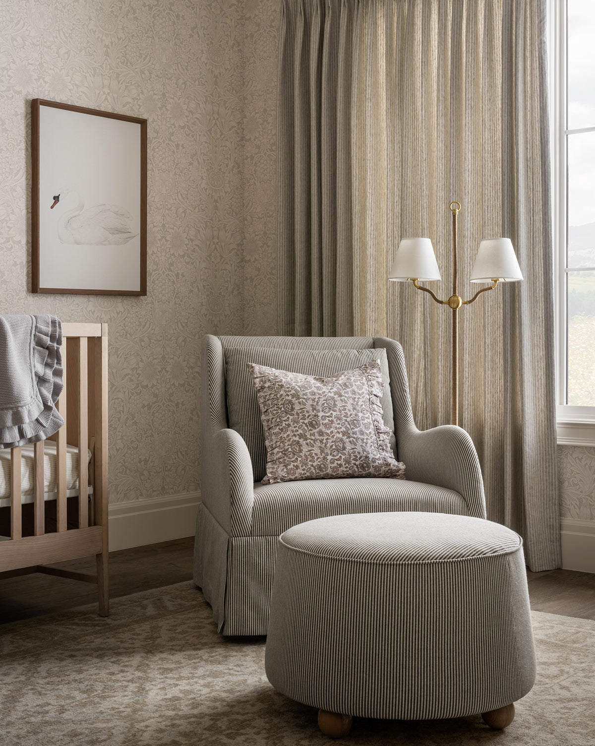 A cozy nursery with a crib, plush seat with the Barnaby Ottoman, floral pillow, dual-shade floor lamp, beige patterned curtains, framed swan art, and softly patterned wallpaper by McGee & Co.