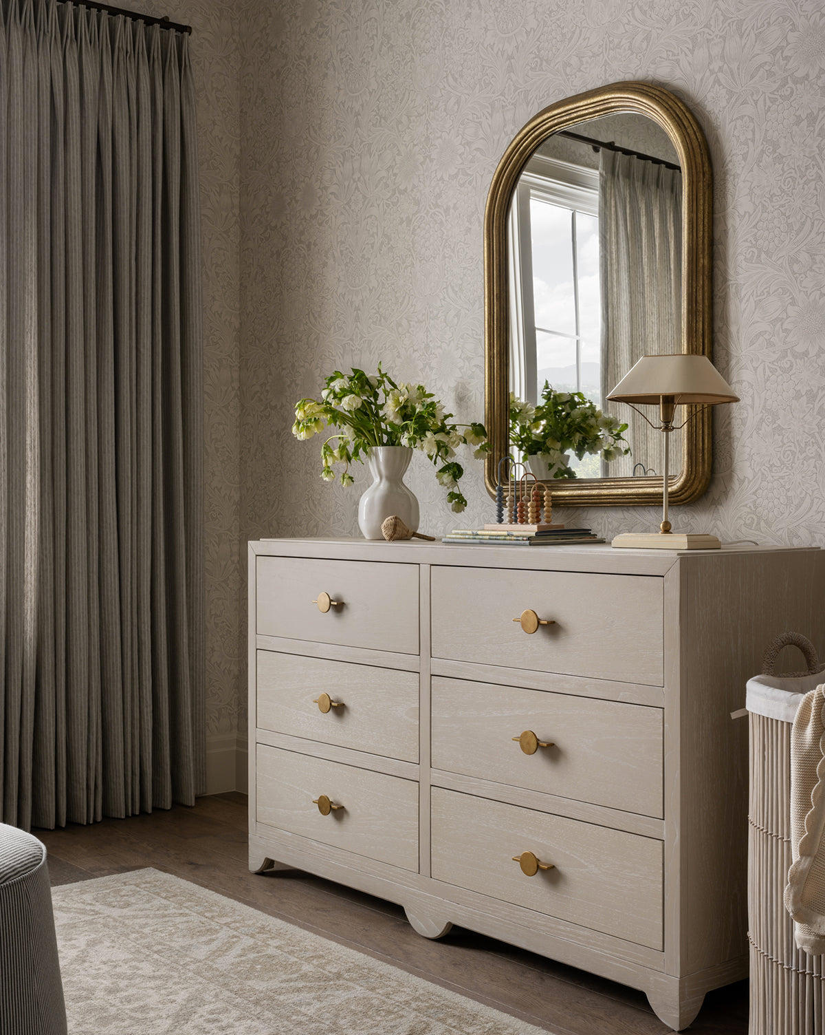 A light wood dresser with round gold knobs stands against patterned wallpaper. On top are a vase, lamp, books, and decor. Above it hangs the Giada Wall Mirror with an elegant moulded frame and antiqued finish, reflecting gray drapes nearby, by McGee & Co.