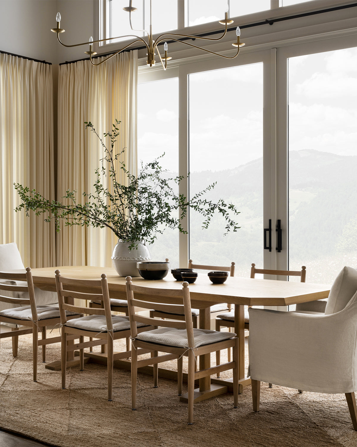A bright dining room showcases the Orville Extension Dining Table with six chairs, a white armchair, rug, and vase of green branches. Floor-to-ceiling windows offer mountain views, complemented by cream curtains and a modern chandelier by McGee & Co.