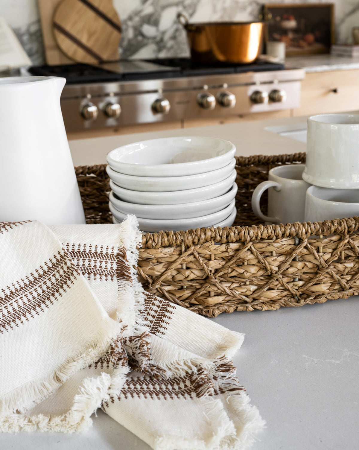 A woven basket holds stacked white bowls and mugs on a kitchen counter. In the foreground, a Woven Stripe Tea Towel with brown stitching and fringe is displayed, with a stove and copper pot visible in the background, by McGee & Co.