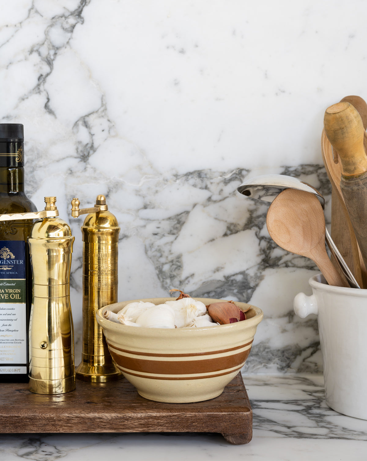 Bottles of olive oil, brass salt and pepper grinders, a garlic bowl, and a ceramic pot with wooden utensils are arranged on a Mango Wood Rectangular Pedestal against a marble backsplash.