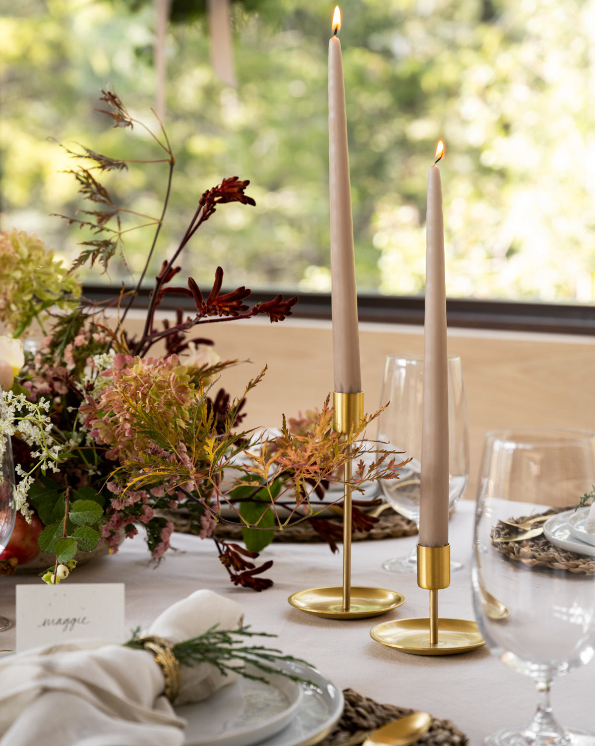 A close-up of an elegant table setting featuring two tall, lit beige taper candles in Brass Taper Holders, surrounded by floral arrangements, glassware, and white plates with beige napkins.