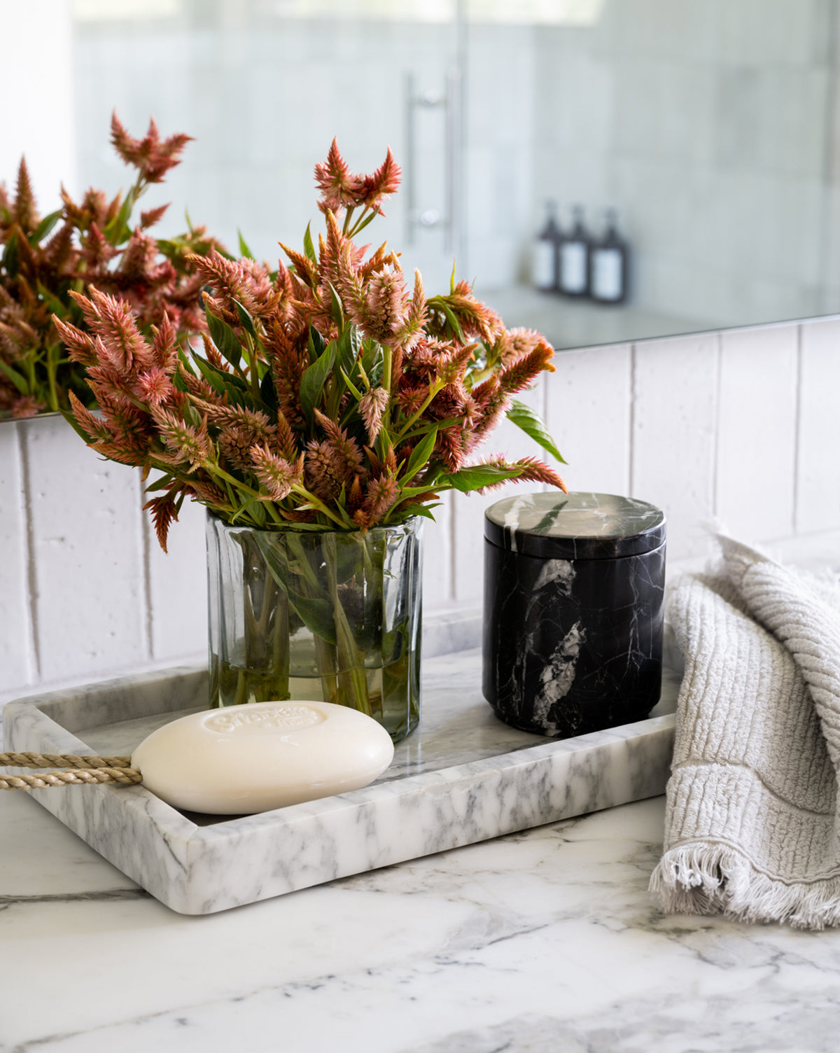 The Marble Tray decorates the bathroom counter, displaying a glass vase with pink-orange flowers, a dark marble candle, soap on a rope, and a folded gray towel. The mirror reflects the calm arrangement.