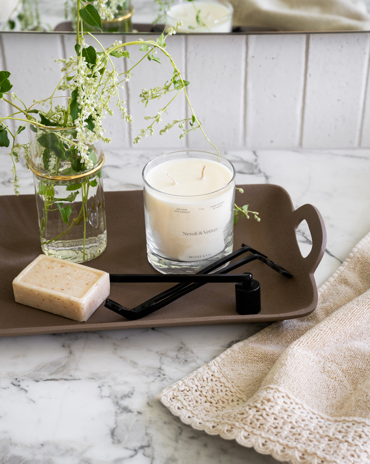 A brown tray on marble holds a glass candle, a Candle Snuffer, wick trimmer, vase with greenery, and soap bar. Next to the tray is a beige lace-trimmed towel. White tiles serve as the background.