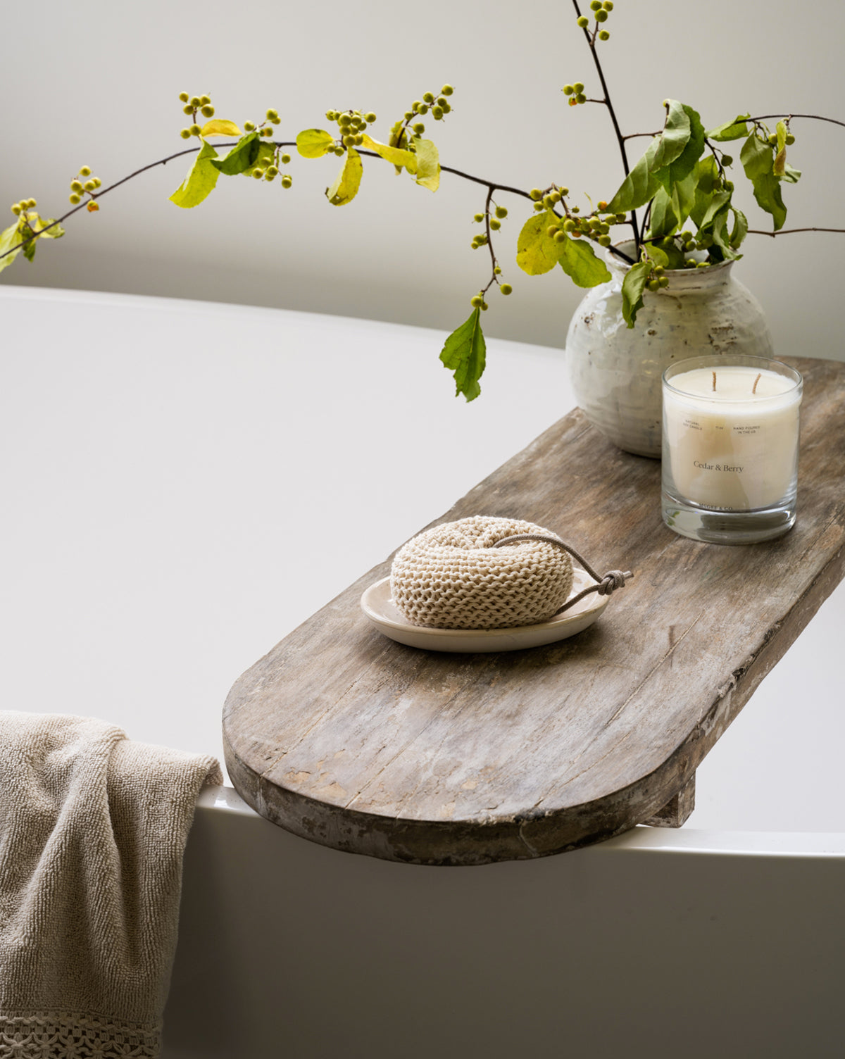 The Rounded Edge Bath Caddy holds a pottery vase with leafy branches, a white candle in a glass jar, and a crocheted scrubber, while a beige towel hangs over the edge of the white bathtub.