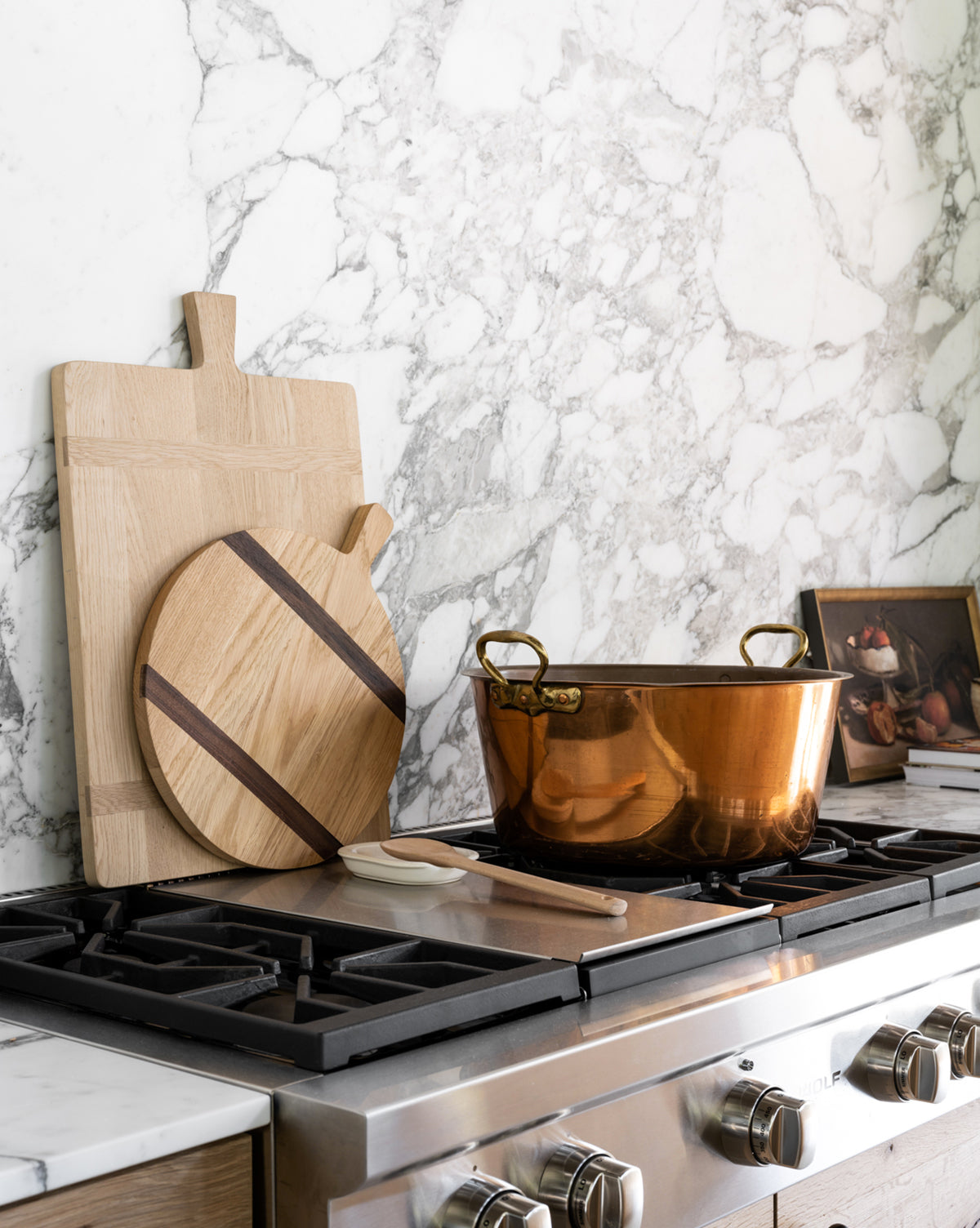 A kitchen stove with a large copper pot, a wooden spoon, two cutting boards—one being the Round Oak Bread Board—and a small framed artwork resting against a white and gray marble backsplash, by McGee & Co.