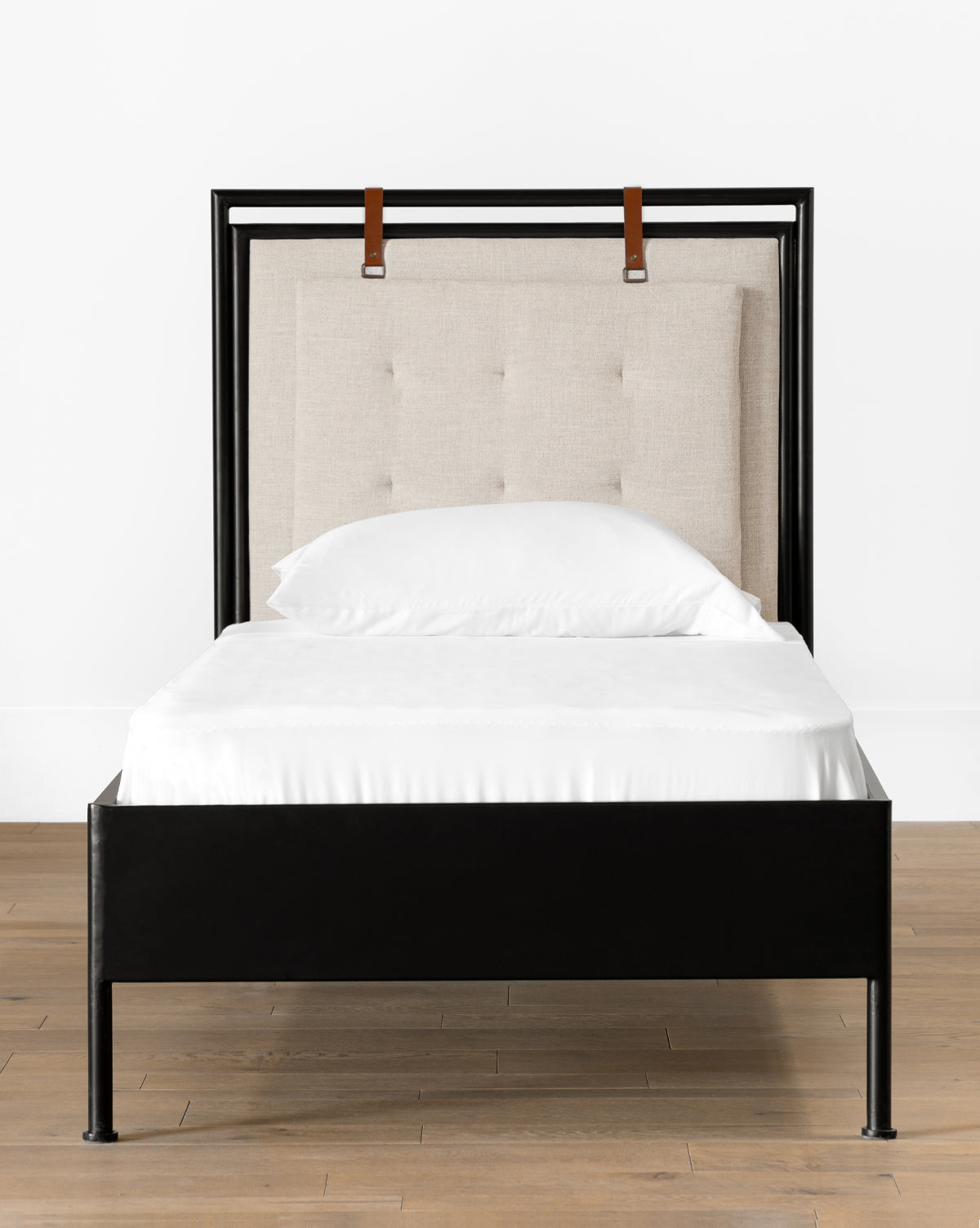 The Sutherland Twin Bed features a black metal frame, padded beige upholstered headboard with button tufting, and two brown leather straps at the top, set on a wooden floor against a white wall, by McGee & Co.