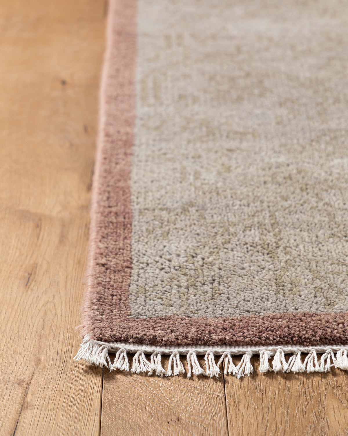 Close-up of the corner of the Suzani Hand-Knotted Wool Rug in light gray with a muted pink border and white fringe, displayed on a wooden floor, by McGee & Co.