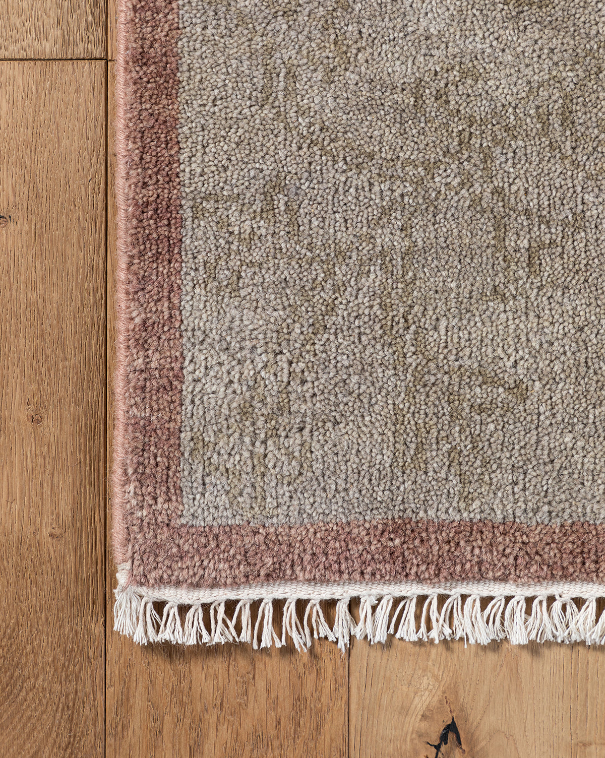 Close-up of the Suzani Hand-Knotted Wool Rug showing a pinkish-brown border, beige center, and white fringe on a wooden floor with visible grain and knots, by McGee & Co.