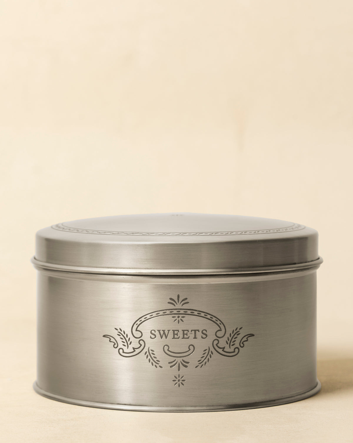 The Sweets Cookie Tin is a round, silver metal tin featuring a decorative pattern and "SWEETS" engraved on the front, adding vintage charm against a plain beige background, by McGee & Co.