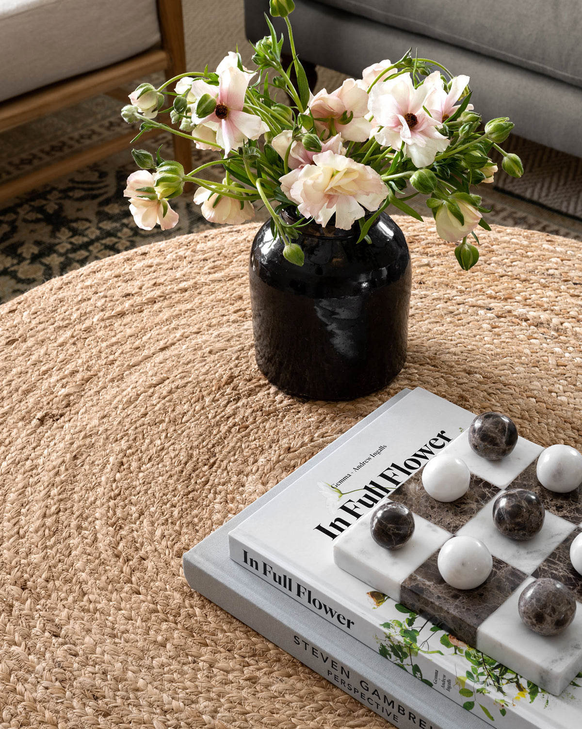 A round woven jute coffee table displays a black vase with pink flowers, two stacked books (including In Full Flower), and the Marble Tic-Tac-Toe Board. A nearby sofa and patterned rug complete this inviting decor scene.