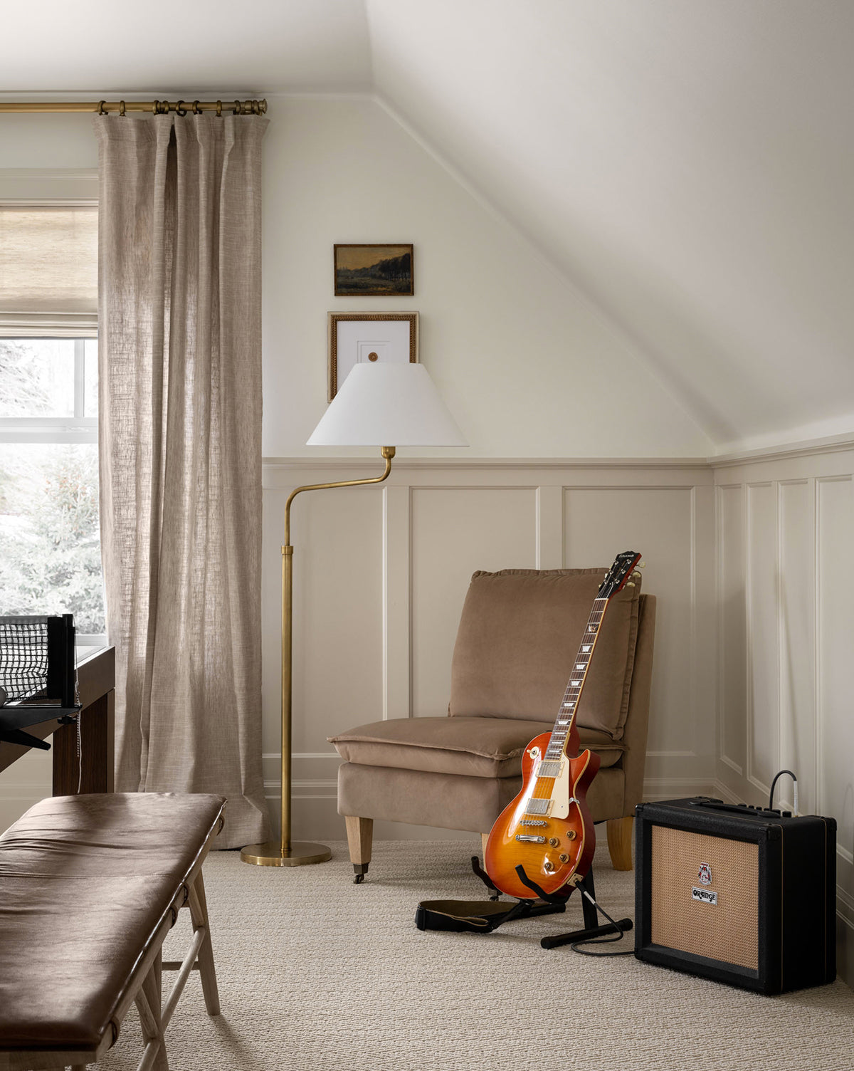 A cozy room with cream walls, a beige chair, the Wainwright Swoop Floor Lamp, electric guitar on a stand, small amplifier, bench, desk, and framed art. Light streams in through a window with sheer curtains, by McGee & Co.
