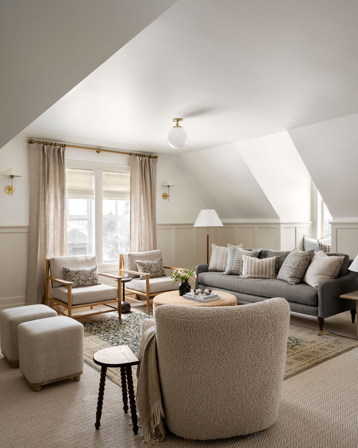 A cozy, neutral-toned living room with a gray sofa, light armchairs, soft rug, round table, ottomans, and an Alice Wood Stool by the large windows with beige curtains under sloped ceilings, by McGee & Co.
