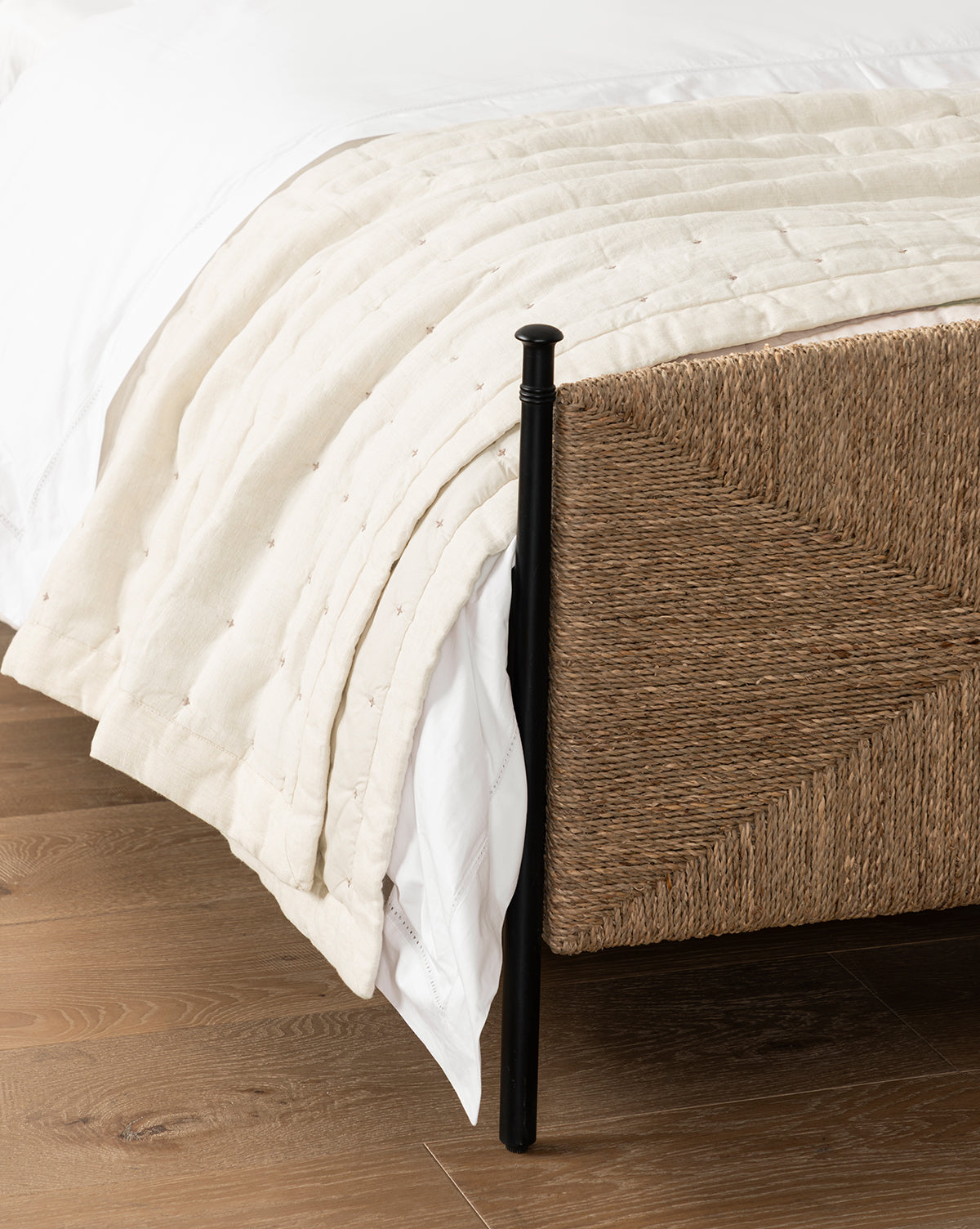A close-up of a bed’s woven rattan frame, white bedding, and the cream-colored cotton Talcott Quilt, all arranged on a wooden floor for a calming neutral palette, by McGee & Co.