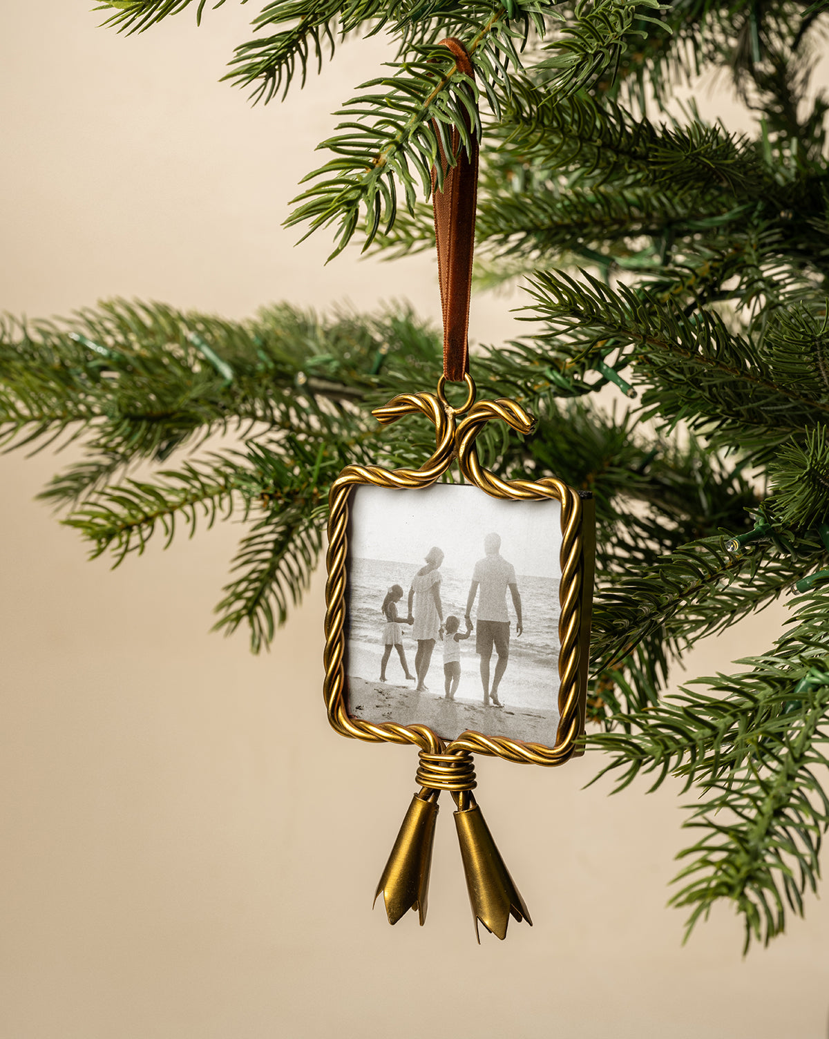 A Tassel Picture Frame Ornament with a gold frame, ribbon, and bells hangs on a Christmas tree branch. Inside is a black-and-white photo of a family of four holding hands, walking along the beach, by McGee & Co.
