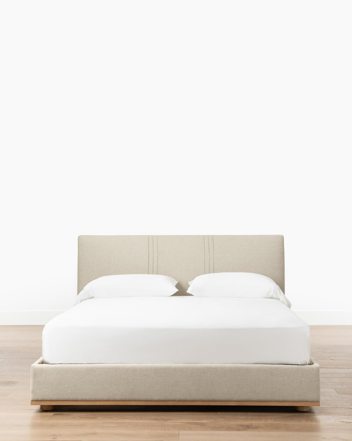 The Taylor Pleated Bed by Makerspalm features a beige upholstered linen frame and minimalist headboard, dressed in white bedding and pillows, placed on a light wood floor against a plain white background—McGee & Co.