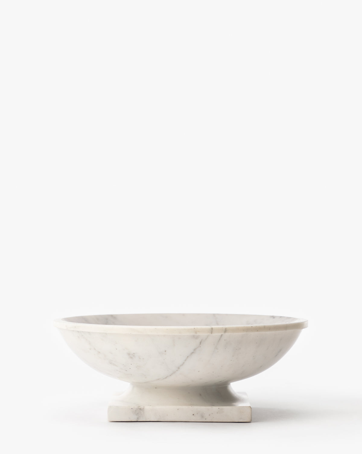 The Tayva Bowl is a sculptural white marble piece with subtle gray veining, featuring a shallow round form, wide rim, and square pedestal base, displayed on a plain white background by McGee & Co.