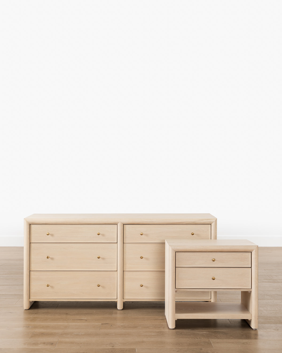 A light wood dresser with six drawers stands beside the Tellerbeck Nightstand, which features two drawers, a simple modern design, and round brass knobs—ideal for effortless bedroom styling against a white wall, by McGee & Co.