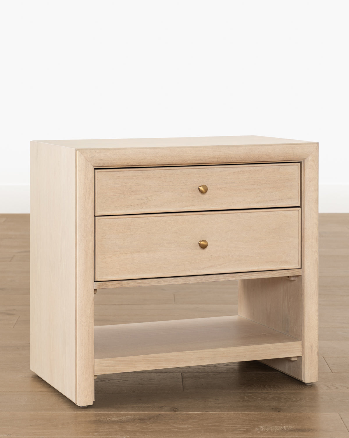 The Tellerbeck Nightstand—a bleached oak piece with two drawers, round brass knobs, and an open lower shelf—stands on a wooden floor against a plain white background, ideal for adding style to any bedroom, by McGee & Co.