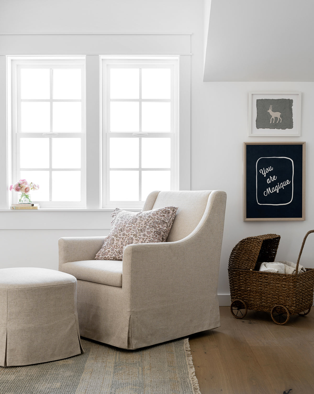 A cozy beige armchair with a matching ottoman sits by a window, adorned with a patterned pillow. A wheeled woven baby bassinet rests nearby. Above, children’s artwork titled “You are Magique” hangs on the white wall over light wood flooring.