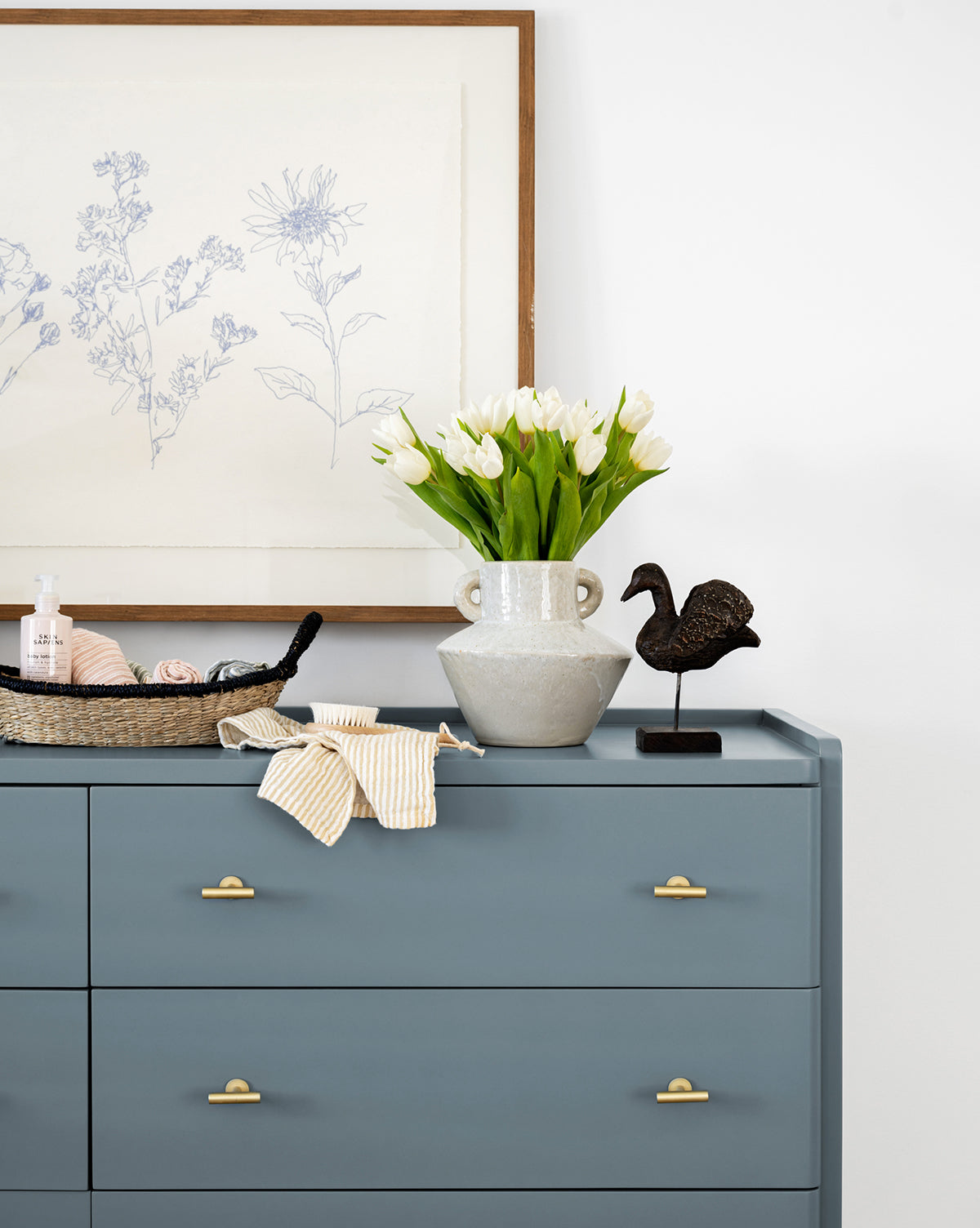 The Petites Fleurs dresser in blue with gold handles displays a vase of white tulips, a woven basket for toiletries, striped fabric accents, and a black bird figurine. A framed botanical print above completes the charming white wall decor.
