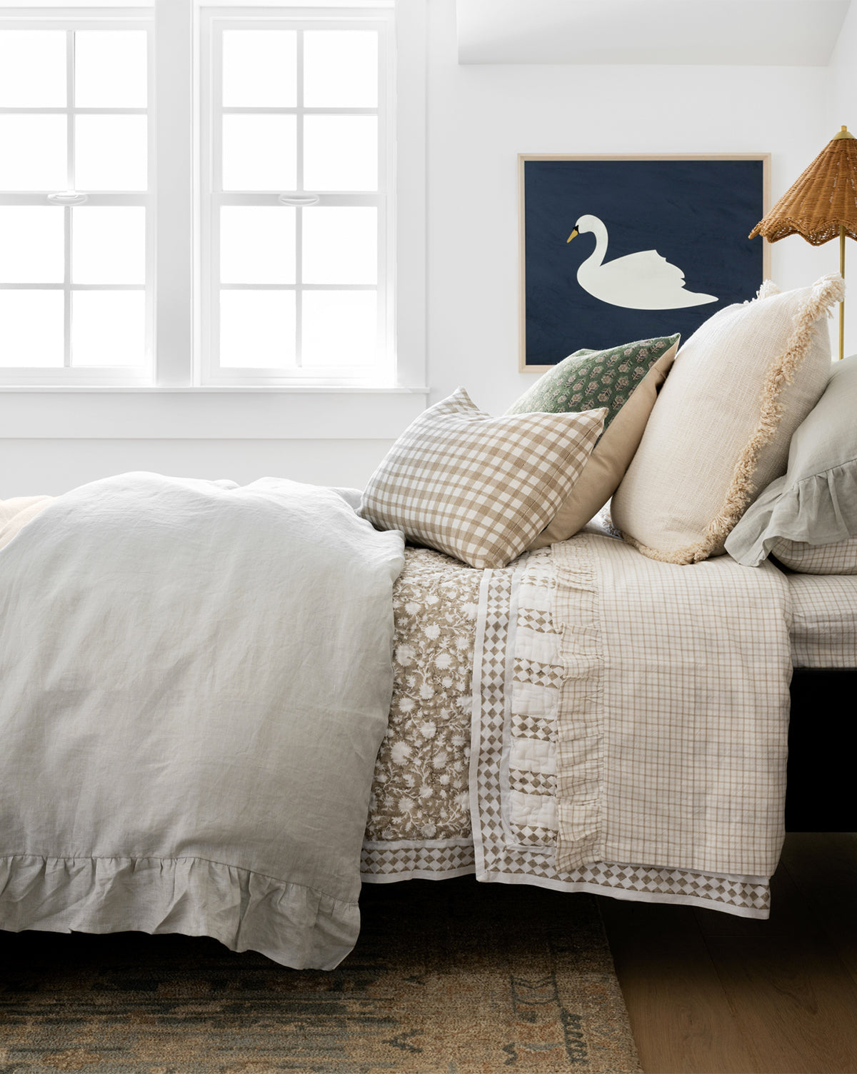 A neatly made bed with light-colored, layered cotton bedding and textured pillows sits by bright windows. Above a bedside lamp with a brown shade, framed swan art hangs; the bed features the soft Nell Ruffled Twin Kids Sheet Set.