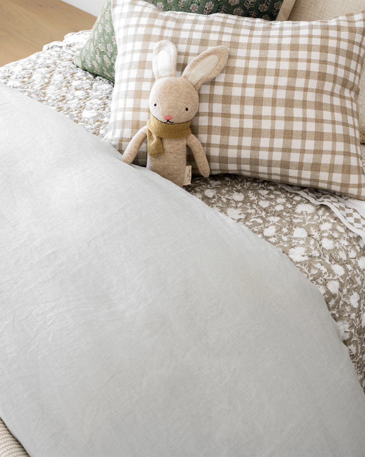 A stuffed bunny with a yellow scarf rests against a beige and white checkered pillow on a bed styled with the Arcadia Block Print Kids Quilt, patterned bedding, and a light gray blanket.