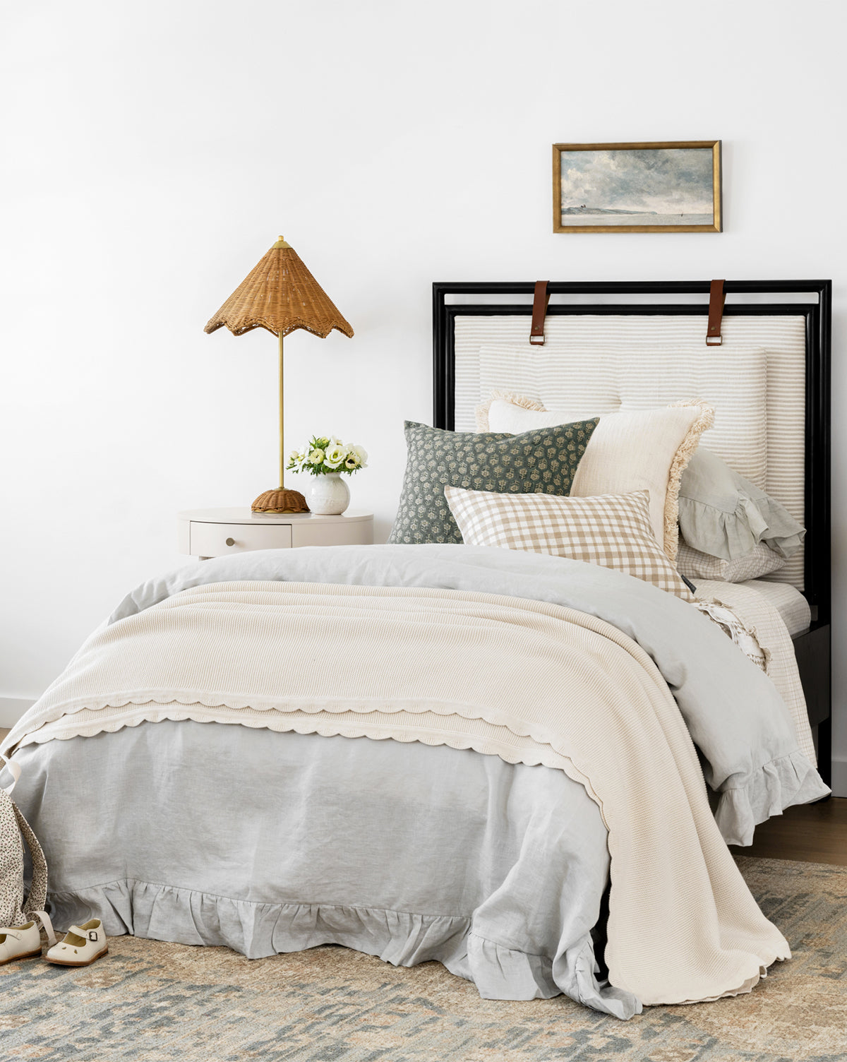 Cozy bedroom with a made bed featuring layered gray and cream bedding, the Pinnacle Knit Kids Blanket (100% cotton), patterned pillows, a bedside table with lamp and flowers, light wall art above the bed, and a soft area rug.