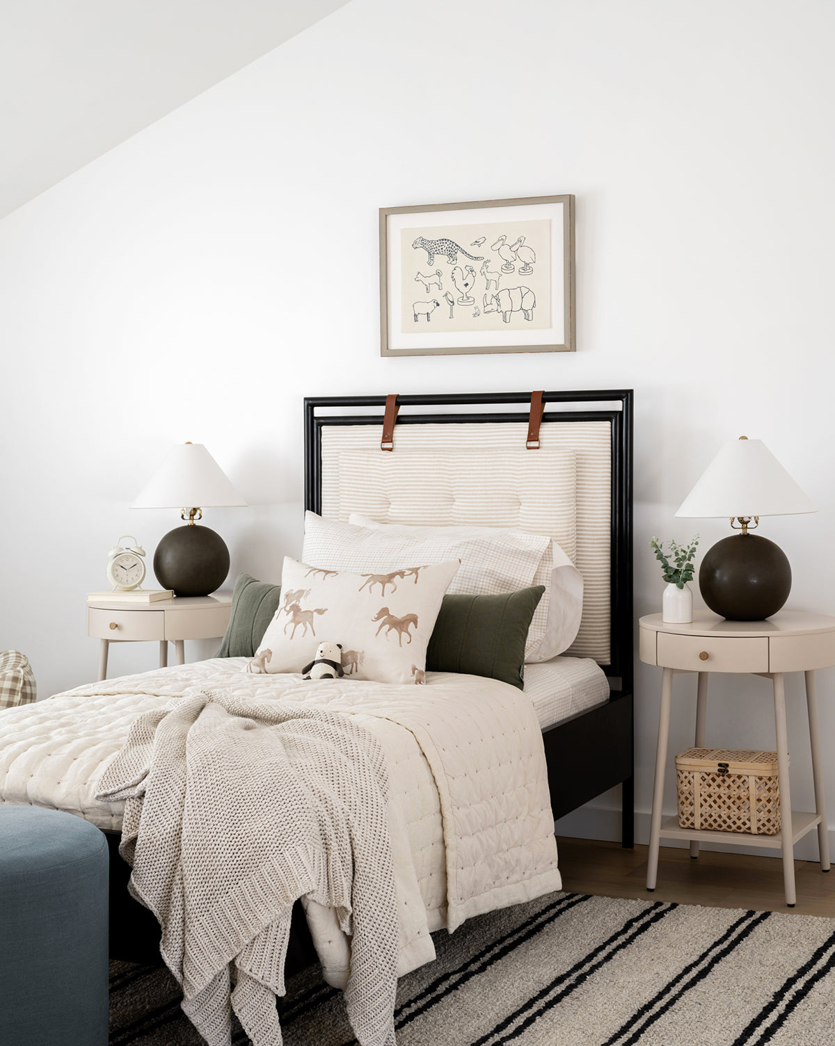 A cozy kids room features a bed with cream bedding, horse-print pillows, and a black headboard. Two round nightstands with lamps flank the bed, and a framed Zoo Cut-Outs print hangs on the white wall above.