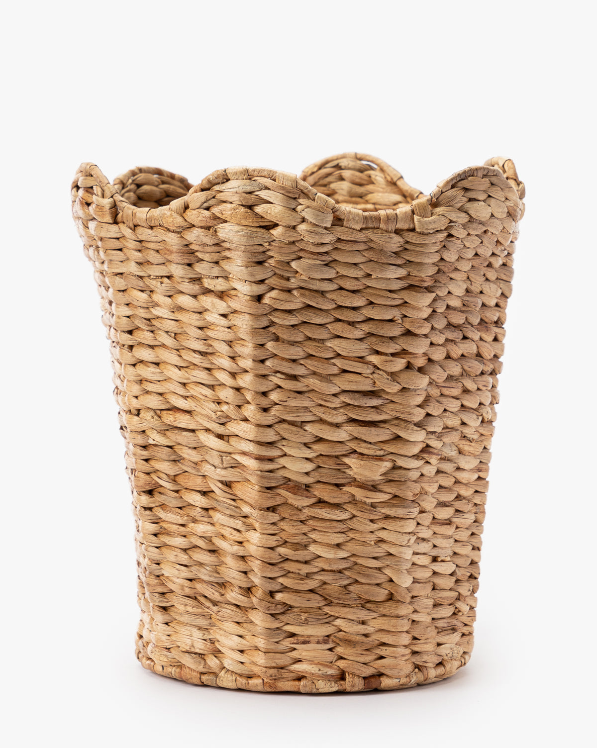 The Tenley Woven Waste Bin features a round, textured surface made from thick, braided water hyacinth fibers in light brown, with a scalloped rim that adds natural charm and elegance. It sits on a white background.