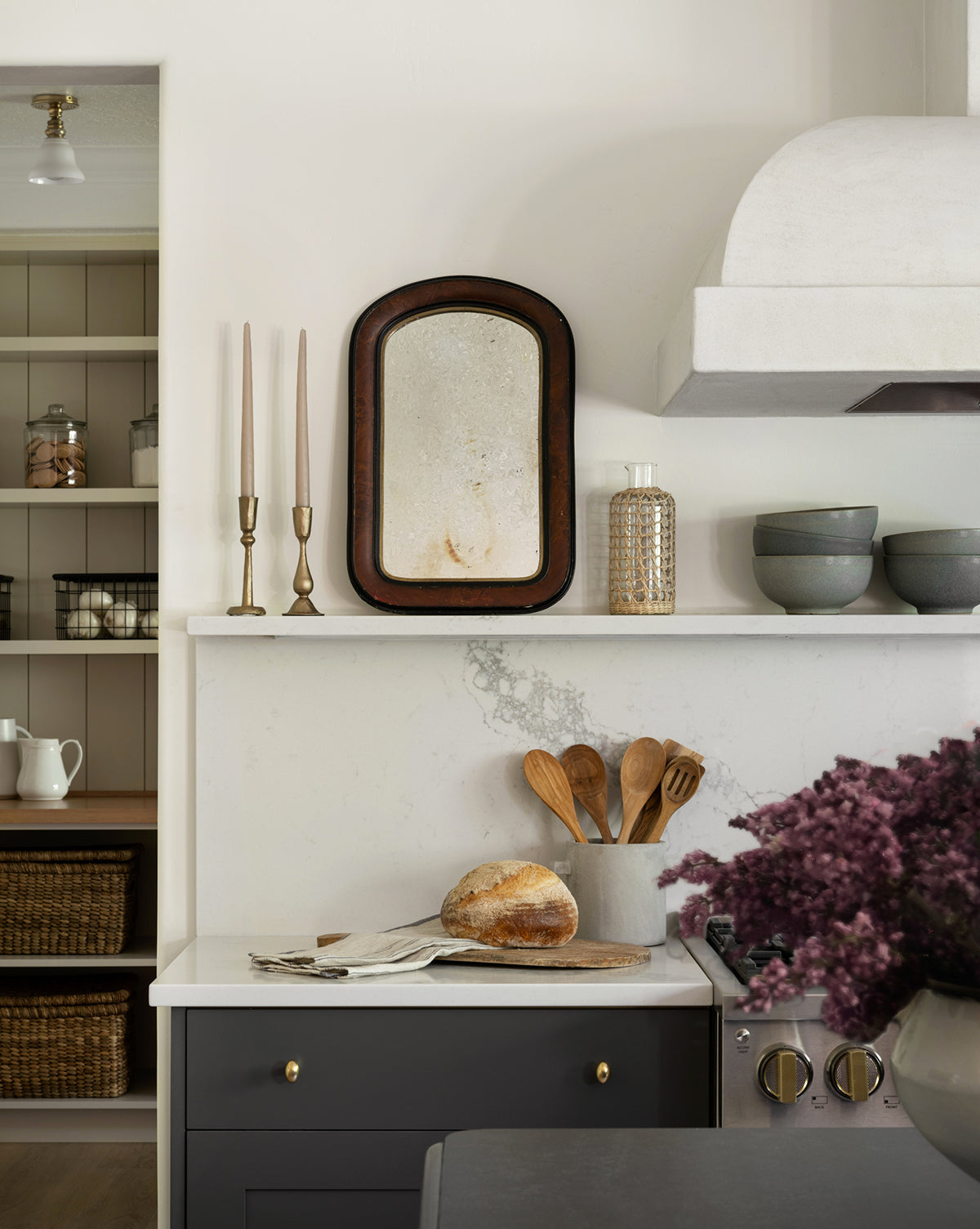 A cozy kitchen countertop features a loaf of bread, wooden utensils in a ceramic holder, stacked bowls, and an Antique Brass Taper Candle Holder. A vintage mirror, purple flowers, open shelves, and baskets complete the inviting scene.