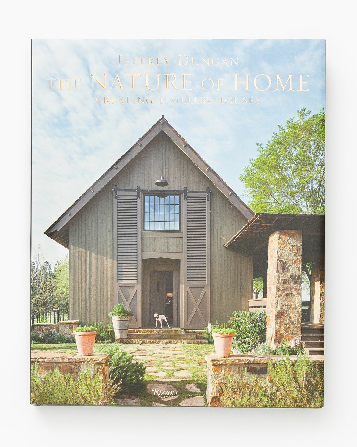 The cover of The Nature of Home shows a rustic wooden house with a dog at the front door, potted plants nearby, and an inviting sense of timeless beauty.