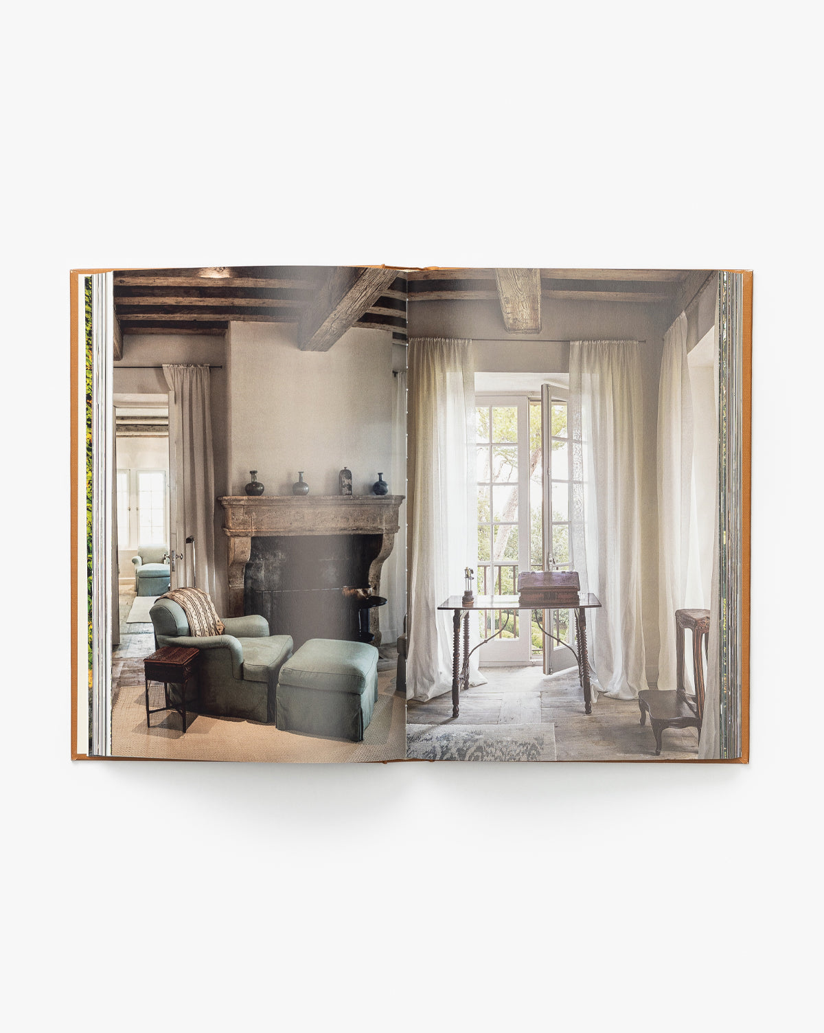 An open design book displays a photo from Three Houses, showcasing a rustic living room with wooden beams, stone fireplace, cushioned armchairs, and large windows—an inviting family home setting.