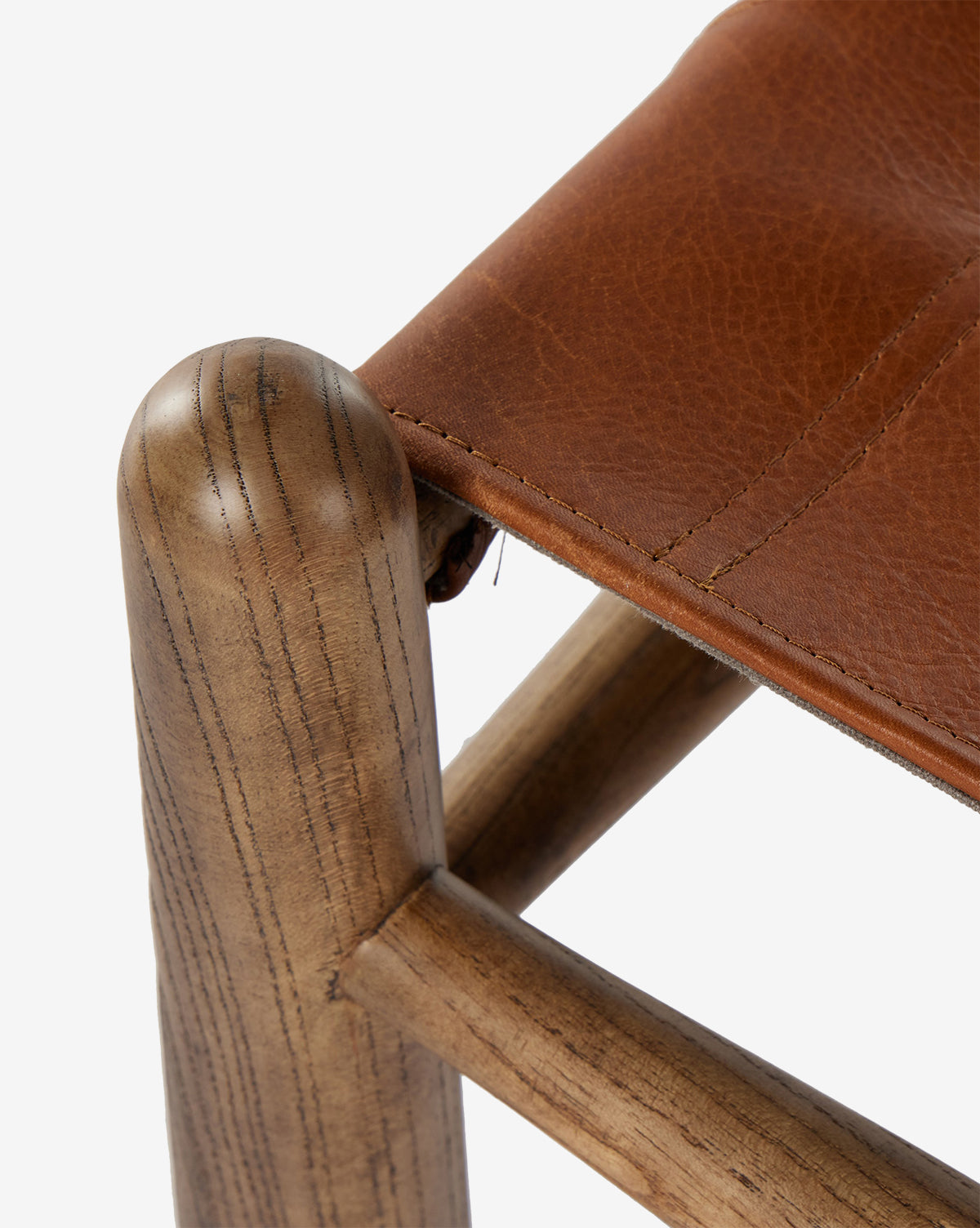 Close-up of the Tierney Ottoman by Four Hands, featuring a solid wood leg with natural grain and smooth finish, topped with a brown stitched top-grain leather seat that is slightly textured. McGee & Co.