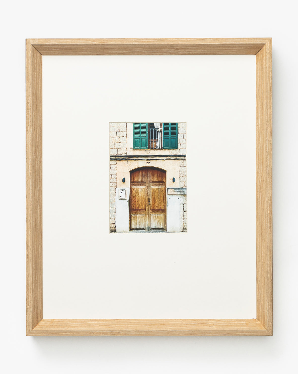 “Town of Deia” is a giclee print depicting a building with a wooden double door, green shutters, and balcony, elegantly presented in a natural woodgrain frame with a wide white mat for modern style.