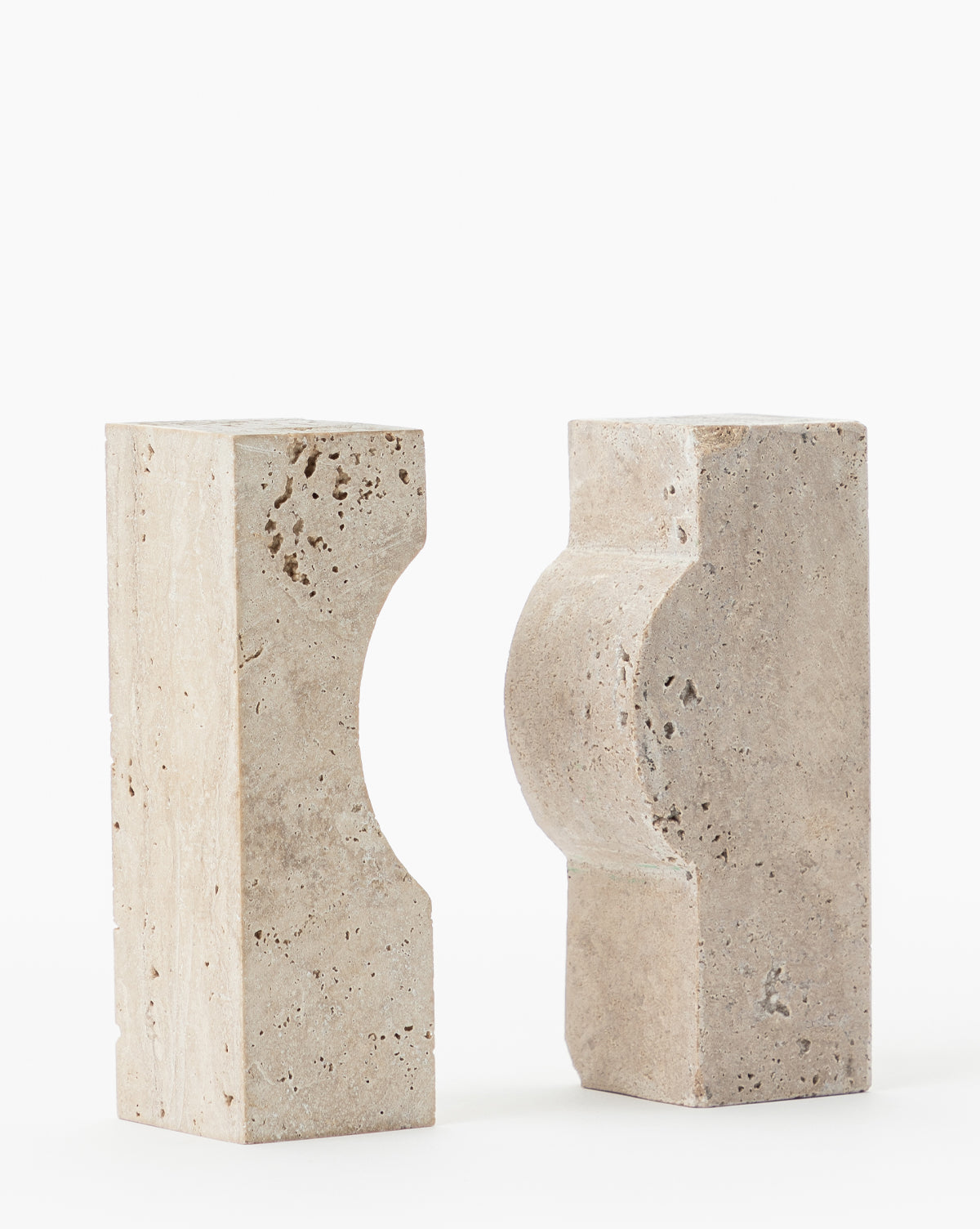 Two beige Travertine Object bookends with geometric cutouts face each other on a white background. The left has a concave curve, the right a convex form, creating a striking complementary decorative accent.