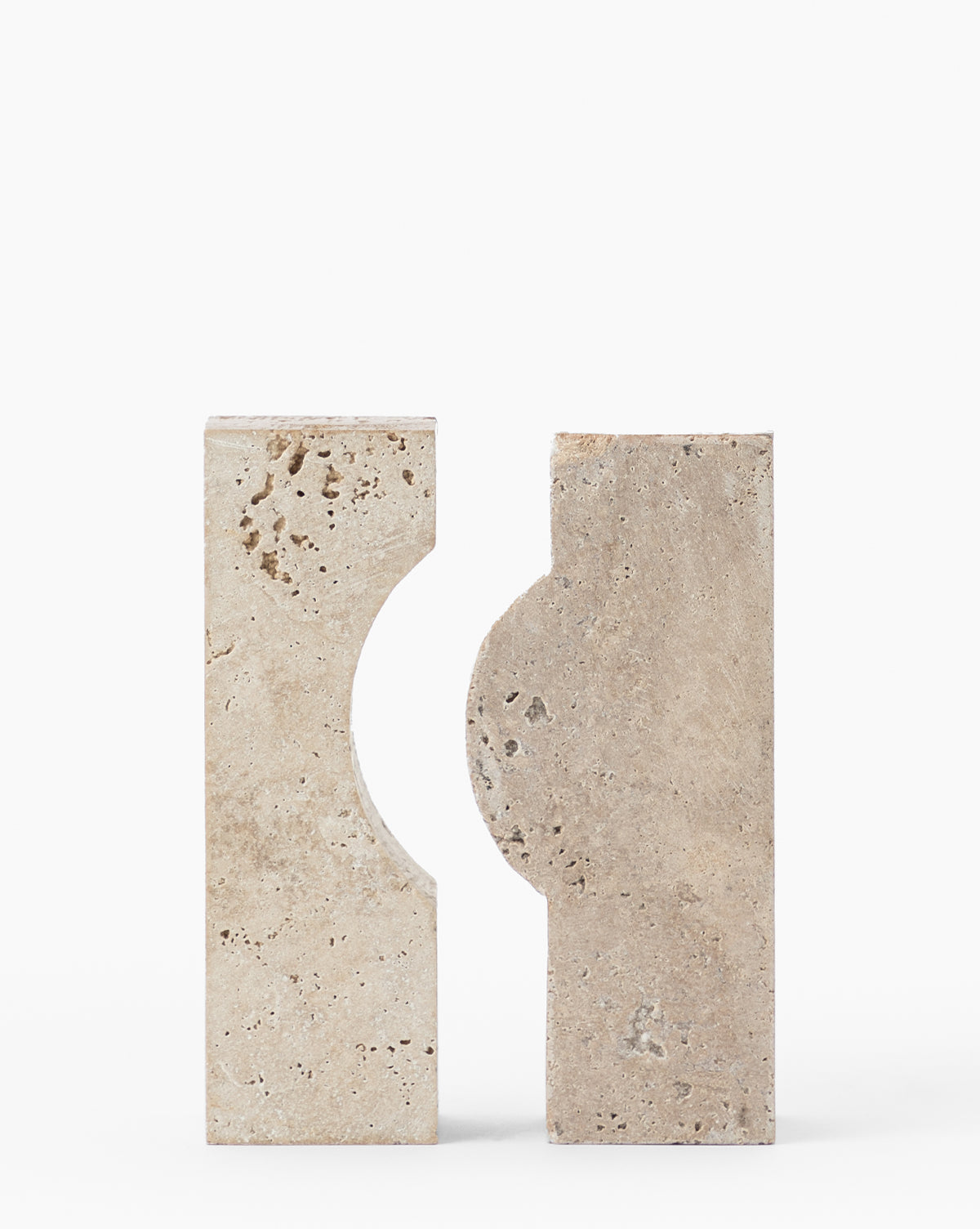 The Travertine Object features two upright rectangular beige blocks of natural travertine, each with a semi-circular cutout on one edge that forms a circular void. The textured surface highlights the stone’s natural character.